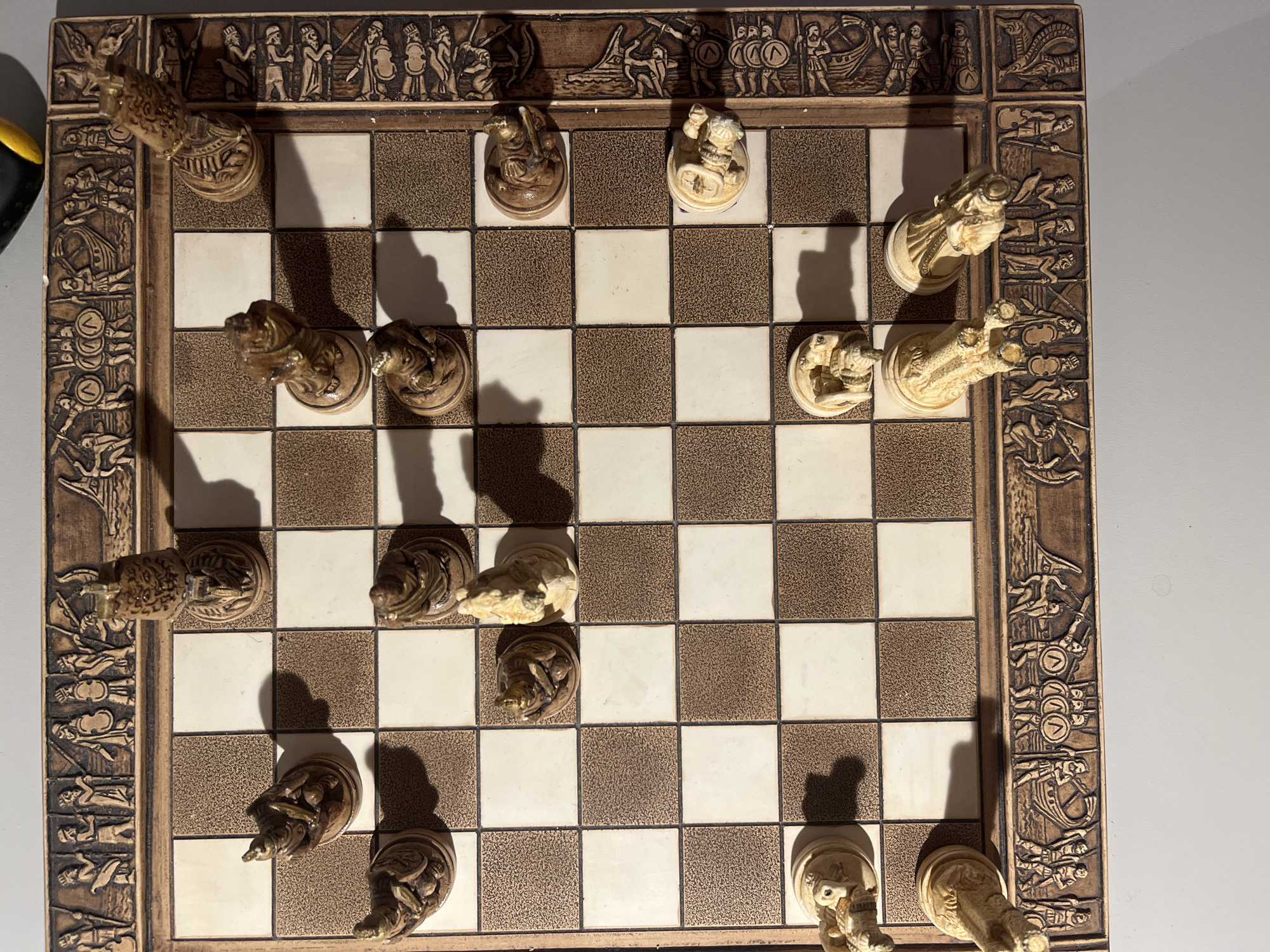 An overhead, slightly angled shot shows a chess game in progress on a carved wooden board. The board is decorated with bas-relief carvings of figures and scenes around its perimeter. Chess pieces, also carved from wood in a classical style, are positioned on the board. Some pieces are upright, while others are lying on their sides, indicating they have been captured. The lighting suggests an indoor setting, casting shadows from the pieces onto the board. The image does not contain any information about the city of Minsk, Belarus, or any specific geographical location.