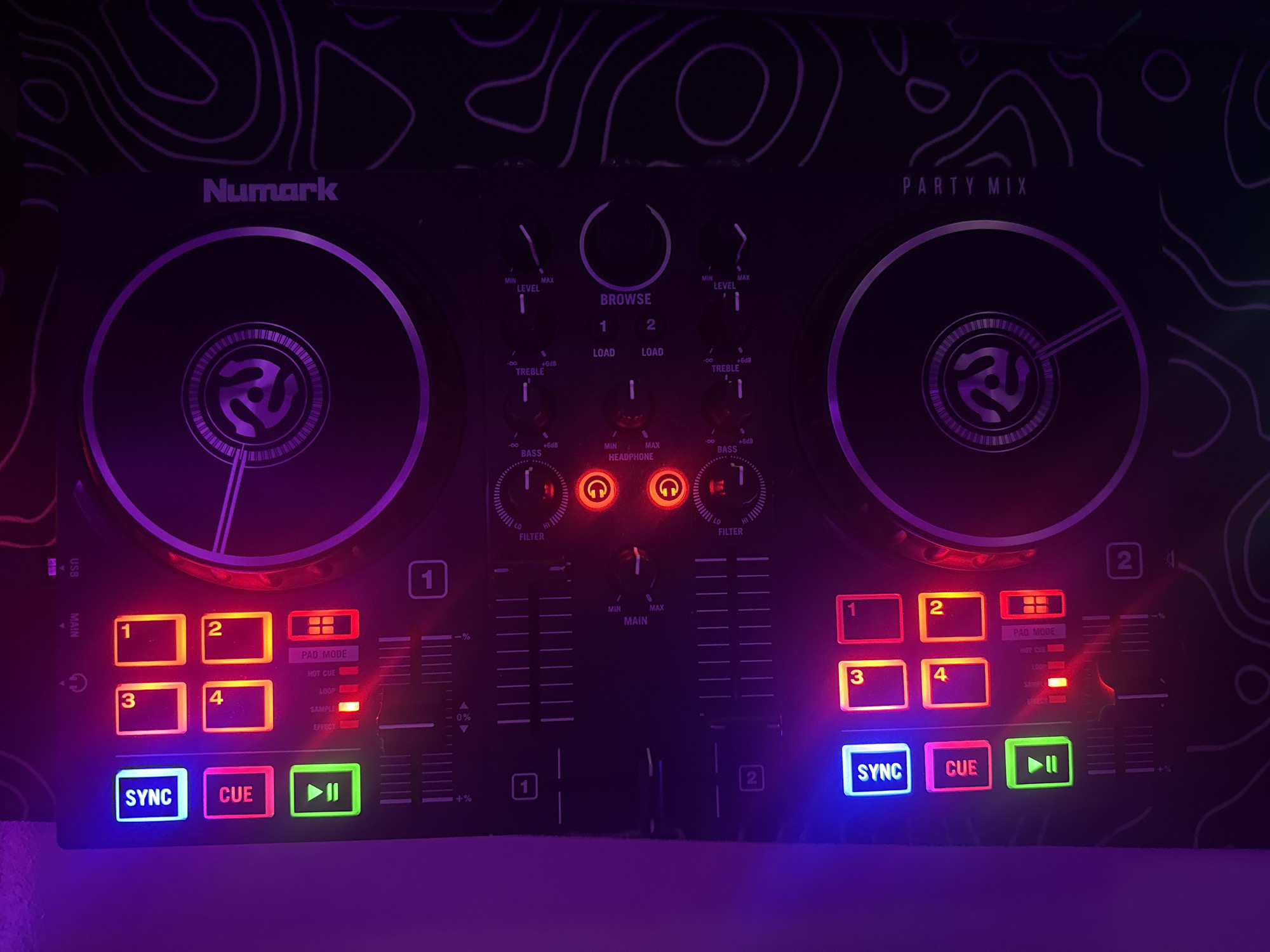 The image shows a Numark Party Mix DJ controller, illuminated by colored LEDs. The controller is positioned on a surface with a topographic map-like pattern. The device features two turntables, several buttons, and sliders, including illuminated "SYNC," "CUE," and numbered pads. The lighting is predominantly purple and pink, casting a glow on the equipment. The orientation of the image is vertical. No specific location in Burgas, Bulgaria, is identifiable from the visual information provided. The scene depicts a static object, the DJ controller, in what appears to be a dimly lit environment.