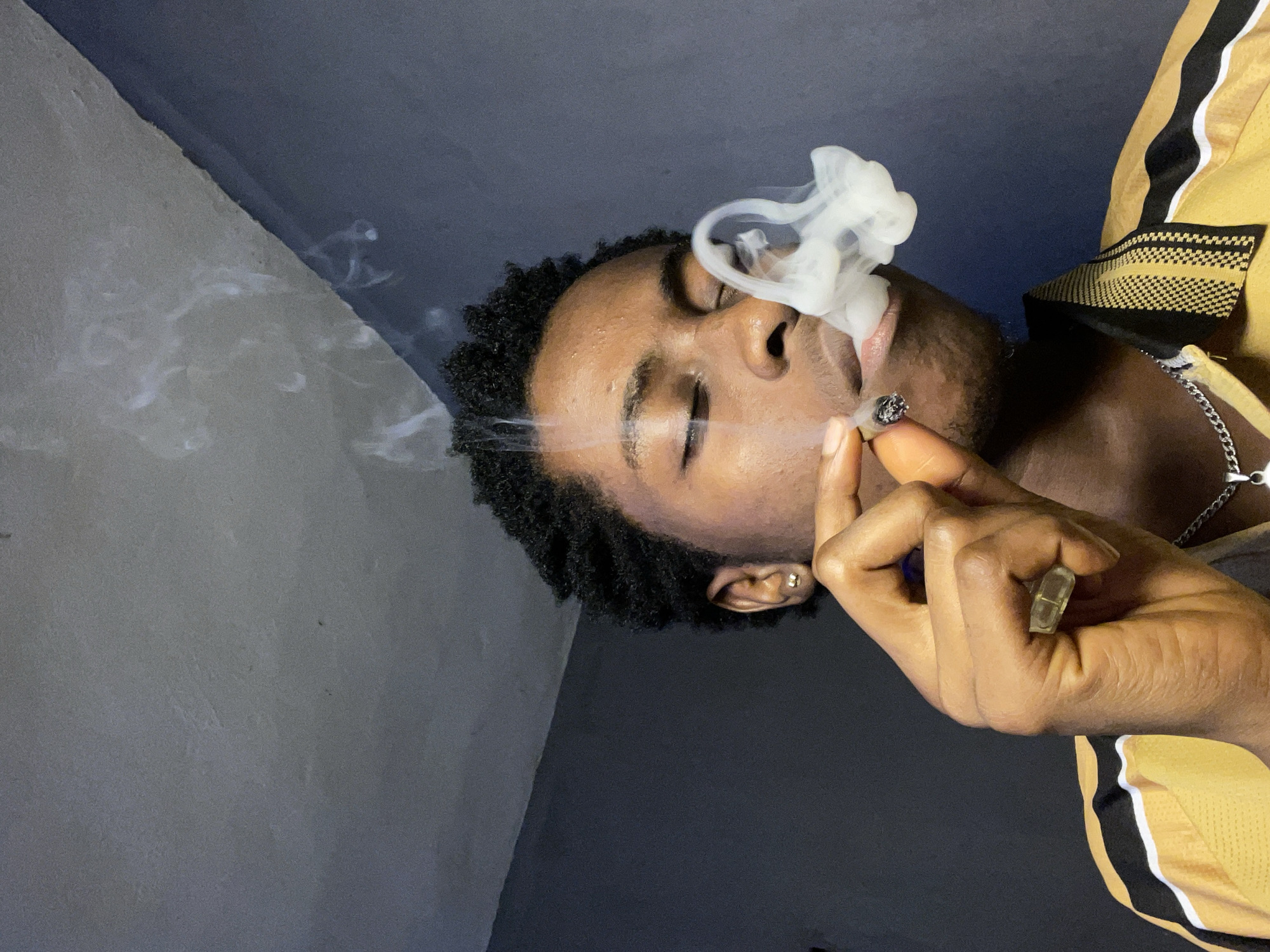 A medium shot depicts a Black man indoors, possibly in Nkpor, Nigeria, based on local context. He has dark, short, curly hair and closed eyes. He is holding and smoking what appears to be a cigarette or joint, exhaling a puff of white smoke. He is wearing a yellow jacket with black stripes and a silver chain necklace. His skin is brown, and he is wearing a gold ring on his right hand and a gold earring in his left ear. The background is a combination of light gray wall and dark blue ceiling or wall.