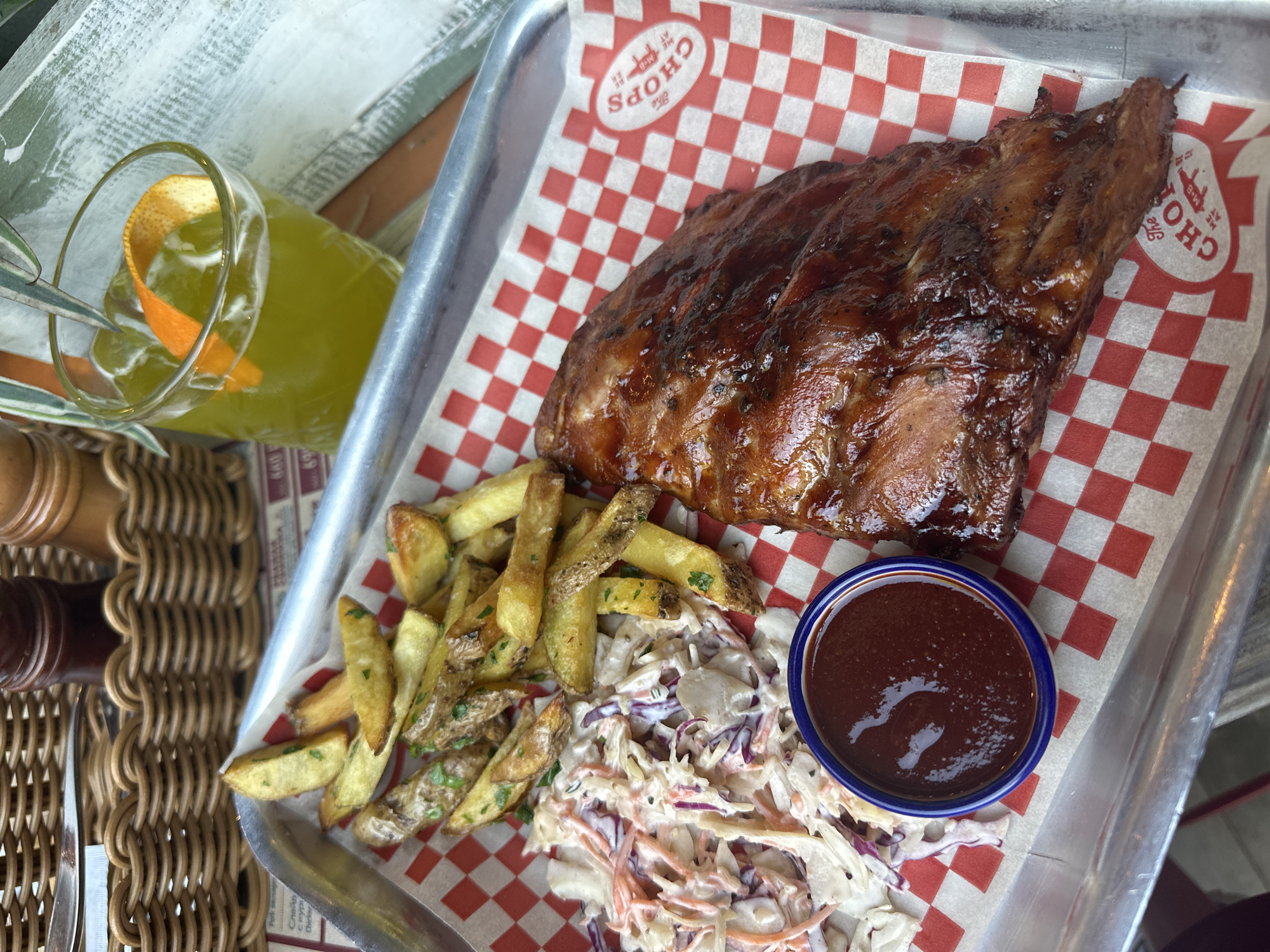 A still, high-angle shot depicts a metal serving tray resting on what appears to be a textured surface. On the tray is a serving of ribs with a glossy, dark brown glaze, alongside a portion of french fries sprinkled with green herbs, and a helping of coleslaw. A small bowl containing a dark sauce, likely BBQ, is also present. The tray is lined with a red and white checkered paper. In the background, a glass of a yellow beverage with an orange peel garnish is partially visible. The restaurant name is visible on the paper lining: "The Chops ME AT M-B ER". The location is unspecified and the claim of Novosibirsk, Russia cannot be verified from the image.