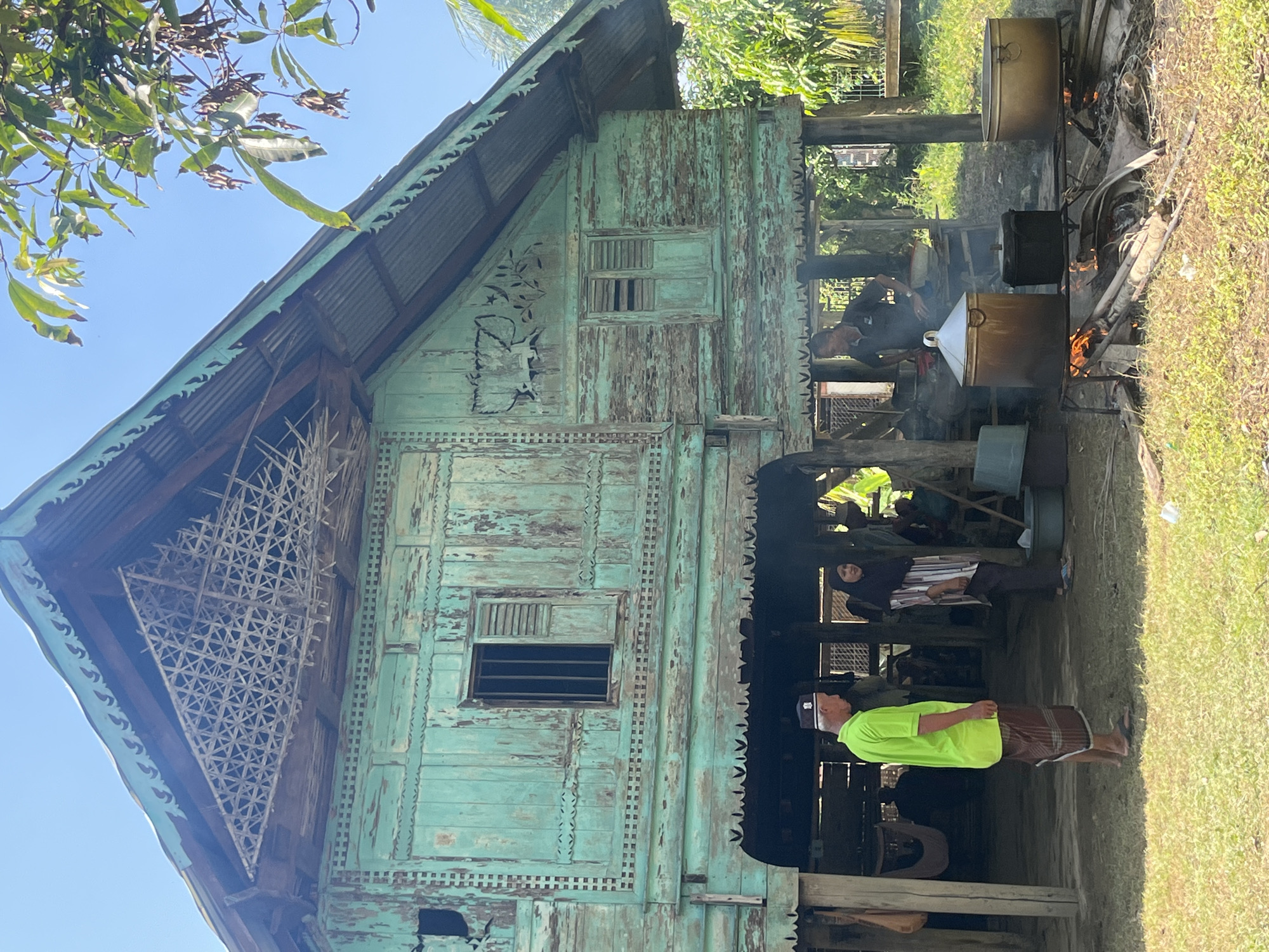 This image depicts an outdoor scene in the village of Pulo, Indonesia, centered around a traditional stilt house with weathered, teal-painted wooden walls. The house features intricate latticework under the gabled roof and decorative carvings on its facade. In the foreground, several large pots are suspended over open fires, suggesting food preparation. A man in a bright green shirt and a sarong stands to the left, facing away from the viewer, near the entrance of the house. To his right, a woman wearing a hijab and a striped top is seen looking towards the cooking area. Another man, partially obscured, is tending to the fires. The ground is covered in grass and dry leaves, and leafy branches of a tree hang over the top right of the frame, casting dappled shadows. The overall atmosphere is that of a rural village setting, possibly during a communal gathering or festival.