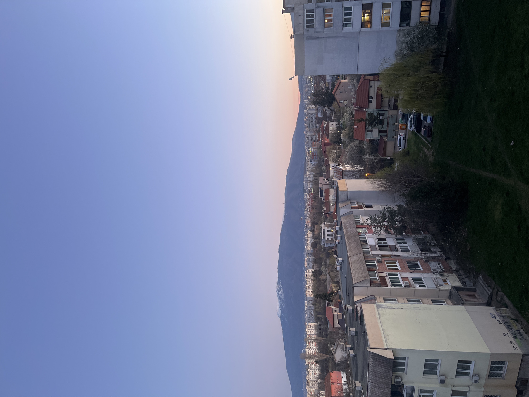 The image captures a view of Sofia, Bulgaria, at dusk, looking towards Vitosha Mountain. In the foreground, there are residential buildings with varied heights and architectural styles. Some buildings show signs of age, including faded paint and external air conditioning units. The mountain range is visible in the middle ground with patches of snow still present near the peaks. The sky transitions from light blue to faint orange hues, indicating the time of day. There's some greenery present, with trees dotted between buildings and some grassy areas visible. Several parked cars and trees line the adjacent street.