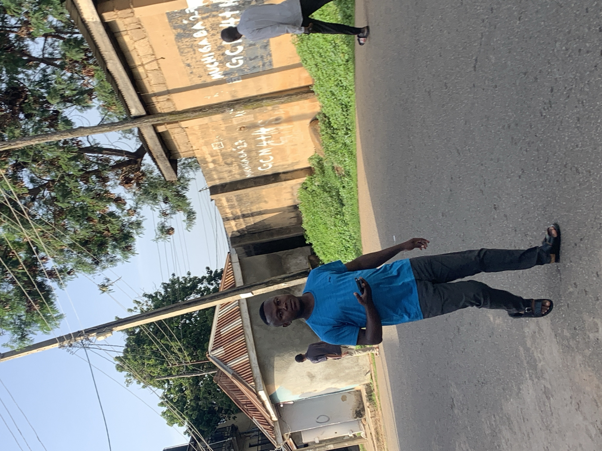 The image depicts a street scene in Jos, Nigeria. A man in a blue t-shirt and dark pants is walking on the road, holding a mobile phone. To his left, a small, weathered building with a corrugated metal roof stands adjacent to a concrete wall. The wall has graffiti on it, including what appears to be writing in white paint. Further down the road, another man in a light-colored shirt and dark pants is walking away from the camera, and a third person is partially visible near the building on the left. Overhead, power lines are strung between wooden utility poles, with lush green trees bordering the scene. The sky is clear and bright, suggesting daytime. The environment appears to be a mixed urban and residential area.