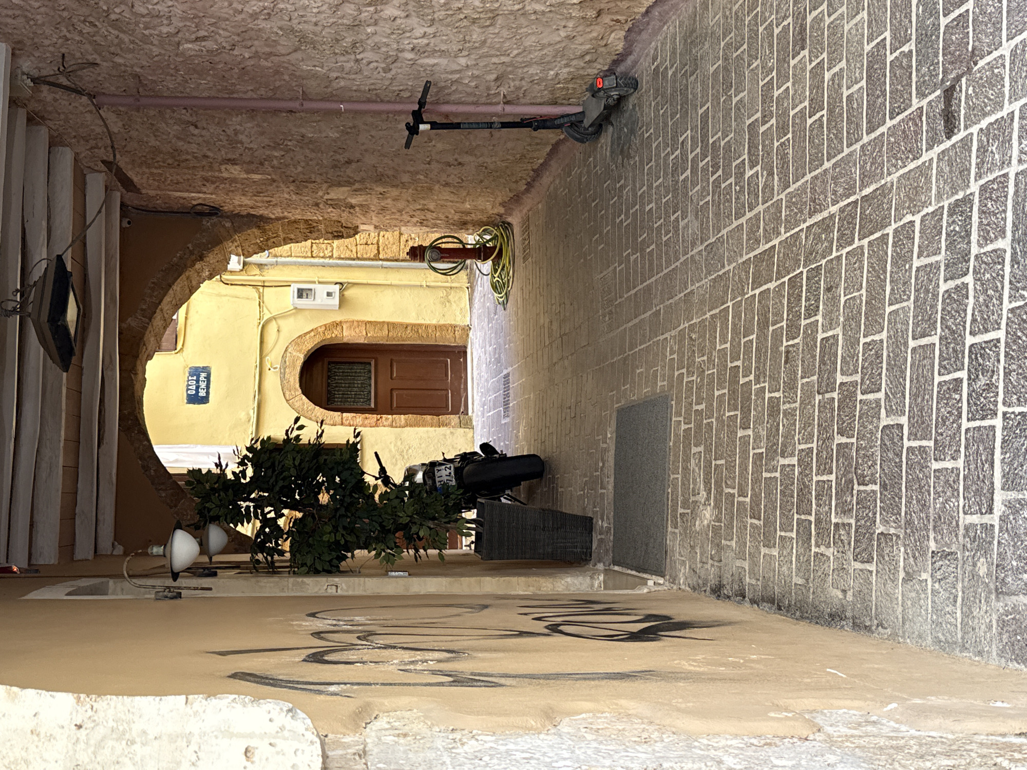 The image depicts a narrow, cobblestone alleyway in Chania, Greece. The alley is flanked by stone walls, one of which is painted a pale yellow and covered in graffiti. The other wall is rough-hewn stone. An arched entryway is visible in the distance, leading to a yellow building with a wooden door. A street sign with Greek lettering, reading "ΟΔΟΣ ΒΕΝΕΡΗ" (Odos Venere - Venere Street), is mounted above the entryway.
In the foreground, to the left, is a black motorcycle parked next to a dark planter with a leafy green tree. To the right, a red fire hydrant with coiled green hoses is situated. Further down the alley, an electric scooter stands upright against the stone wall. The cobblestone pavement is laid in a diagonal pattern with a contrasting strip running down the center. Overhead, exposed wooden beams support a section of the alley. A street lamp with two globes is attached to the yellow wall on the left.