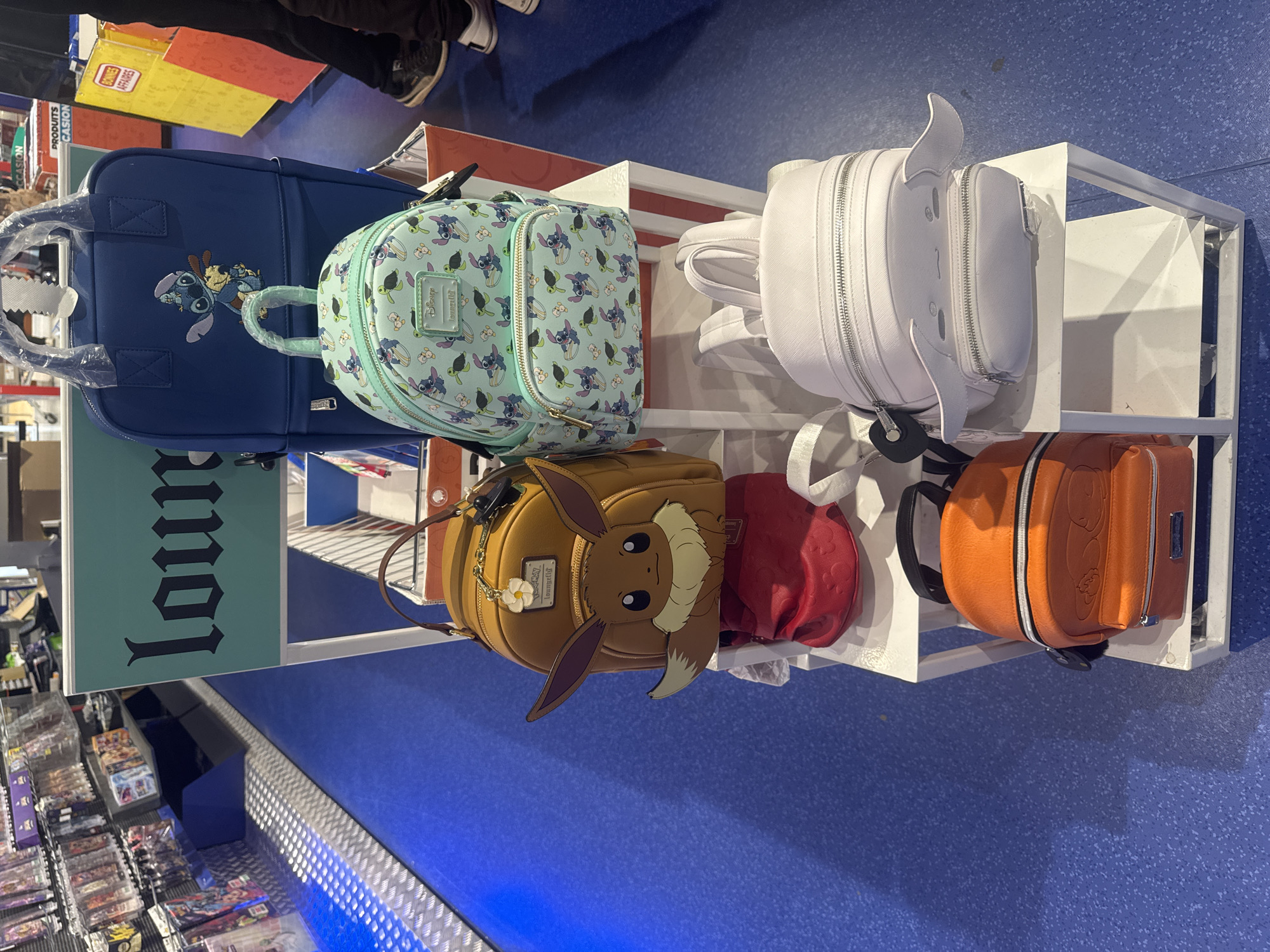 The image shows a retail environment, likely a store, featuring a display of character-themed backpacks. The backpacks are arranged on white shelving units. In the foreground, on the lower shelf, is an orange backpack with an embossed character design, possibly from the Pokémon franchise. Above it, on the middle shelf, sits a tan backpack with Eevee's face, ears, and tail prominently displayed, along with a small white flower charm and a Loungefly tag. To the right of the Eevee backpack is a white backpack with zipped compartments and what appear to be small, white wings. Further back on the middle shelf, a light blue backpack is adorned with Stitch and other characters from Disney's Lilo & Stitch. To the left of the blue backpack is another, larger blue backpack with a Stitch applique. In the background, shelves are stocked with various items, and a sign with "loun" in stylized lettering is visible. The floor is a speckled blue. The overall impression is a merchandise display, likely in a specialized store catering to fans of anime and pop culture. The question about Leers, France is not directly answerable from the visual information present in the image.