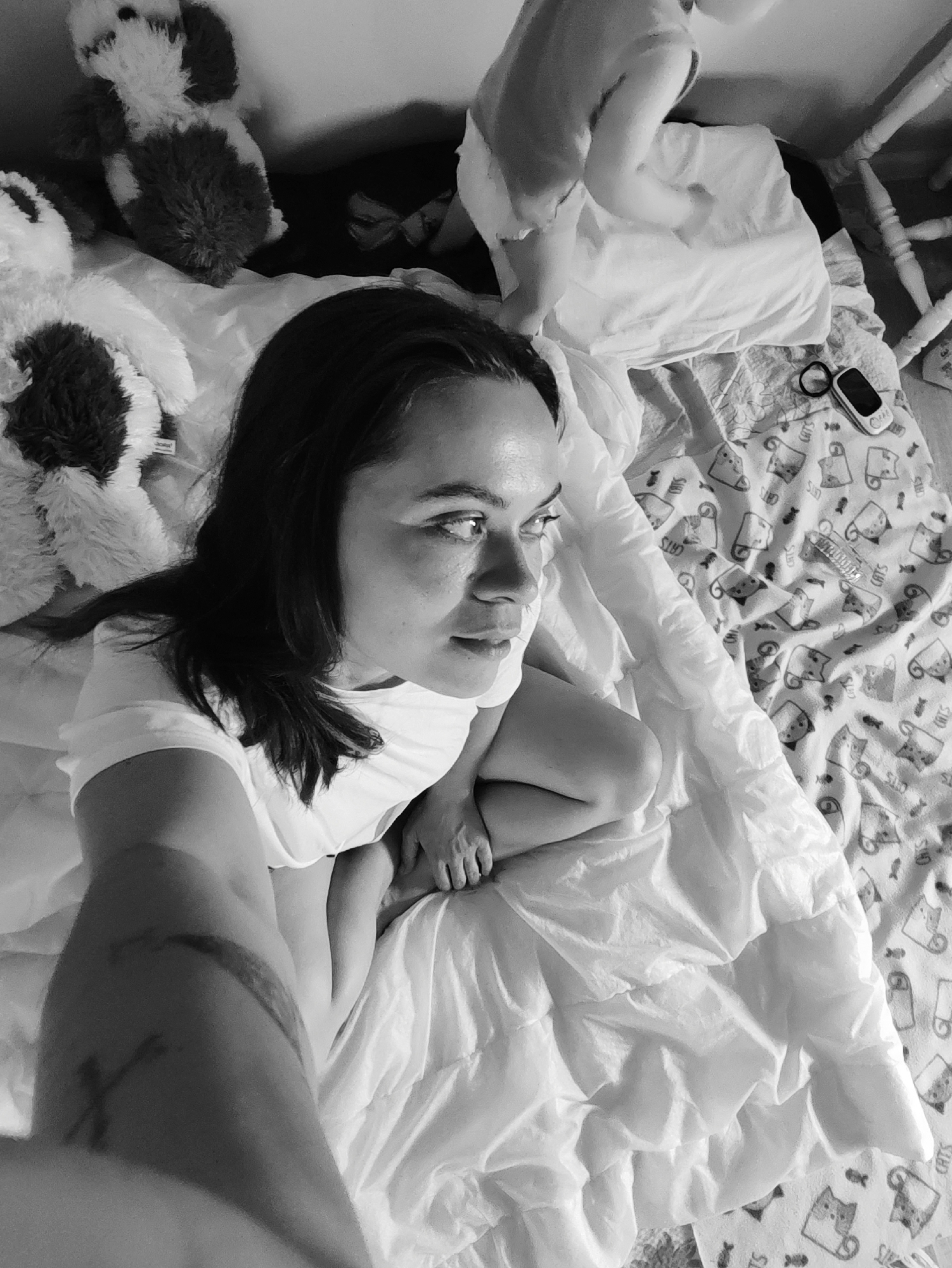 A black and white, high-angle shot shows a woman sitting on a bed. She is wearing a white t-shirt and is looking up and to her right. Her dark hair is parted on the side. Her legs are bent and drawn up to her chest. Her arms are in front of her, and her hands are clasped around her legs. Her left arm, which is angled into the frame, has a tattoo. Next to her there are 2 stuffed animals. Behind her, there is an infant on top of a pillow. To the right of the pillow is a blanket with a pattern of cats and fish bones on it. A hair tie and small electronic device sit on top of the blanket. Part of a white bed frame is visible in the top right corner of the frame. The location is presumed to be a bedroom in Unknown, Unknown.