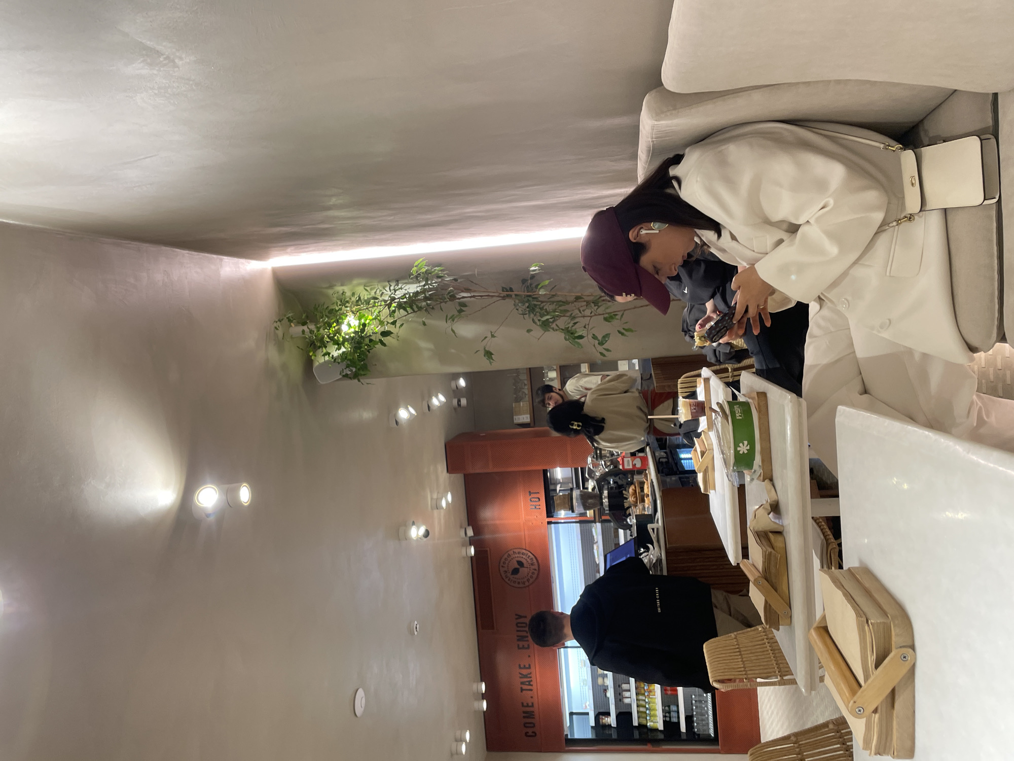 The photo captures the interior of a cafe in Yerevan, Armenia. The foreground features a woman wearing a white coat and a dark red cap. She is holding a smartphone and possibly placing an order. Other customers are seen in the background, including one dressed in beige and another in black facing the order counter. A retail space with the words "COME. TAKE. ENJOY." displayed on a brown wall can be seen. The cafe's interior includes bright lighting, white tables, and woven chairs, adding a minimalist aesthetic. A potted plant is visible on the left side of the image.