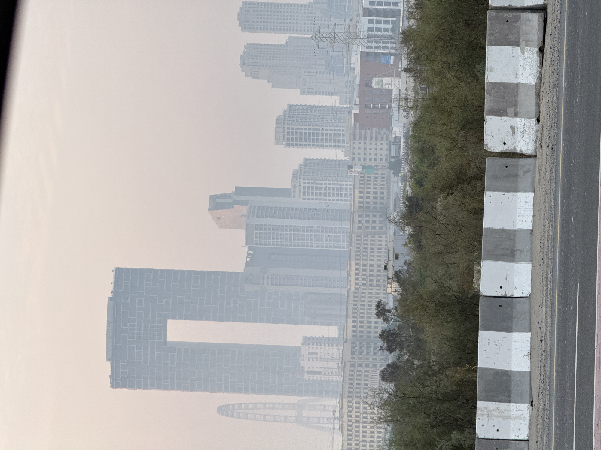 The image shows a hazy cityscape in Dubai, United Arab Emirates, with several skyscrapers dominating the skyline. A distinctive building with a large rectangular opening in the center stands prominently in the foreground. To its left, a Ferris wheel-like structure is partially visible. In the middle ground, a long, low-rise building with numerous windows spans across the scene, backed by a dense collection of residential towers. A power pylon with electrical wires is visible on the right side of the image. In the immediate foreground, a concrete barrier with alternating black and white stripes separates the road from a strip of land with sparse vegetation. The sky is overcast and hazy, obscuring some of the finer details of the distant structures. There are no people visible in the image.