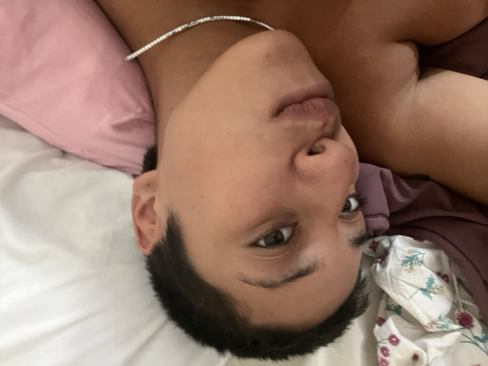 The image displays a single individual in a close-up, rotated 90 degrees clockwise, depicting them lying supine in an indoor setting, presumably a bedroom. The subject, an adult with short, dark hair, has a medium skin tone. A thin, silver-colored necklace is visible around their neck. Their eyes are open and directed straight at the camera, maintaining a neutral facial expression. The upper torso is partially exposed, revealing a shoulder. The individual rests on a white sheet, with a pink pillow positioned above their head (relative to their body orientation). Adjacent to the individual, on the right side of the frame, are bedding materials consisting of a white fabric patterned with green leaves and pink/purple flowers, and a solid purple fabric. No visual cues are present within the image to identify a specific location within Buenos Aires, Argentina.