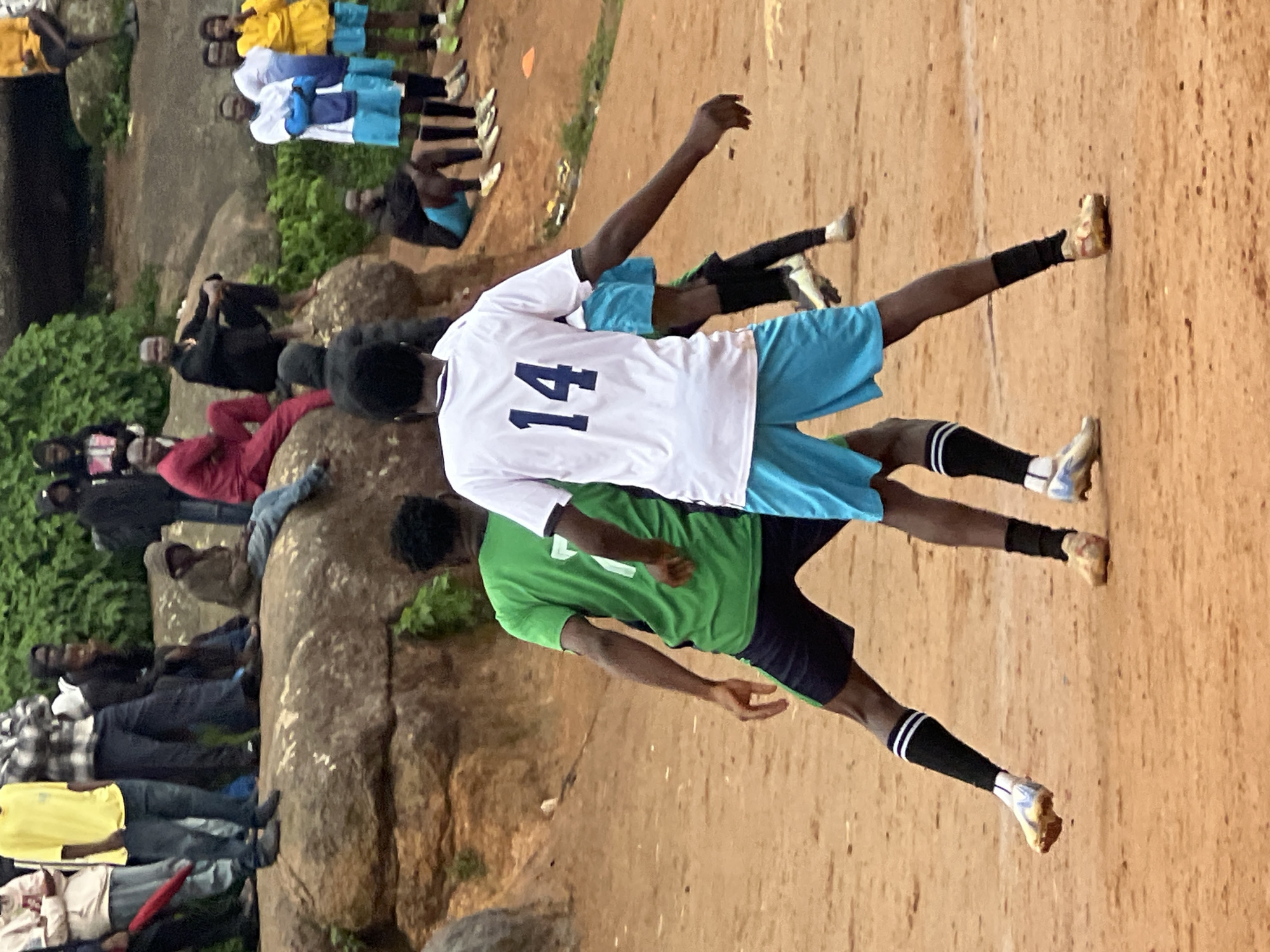 The image depicts a street soccer game on a dirt field in Jos, Nigeria. Two male players are in the foreground, facing away from the camera and towards each other. The player on the left wears a green jersey with the number 7 and dark shorts, while the player on the right wears a white jersey with the number 14 and light blue shorts. Both players have black socks and soccer cleats. Other individuals, some in soccer attire and others in casual clothing, are visible in the background, seated or standing on a large rocky outcrop. The environment is arid, with a reddish-brown dirt ground and scattered green vegetation. The rocky formation suggests a natural landscape within or near the city. The overall scene captures a moment of athletic activity in a community setting.