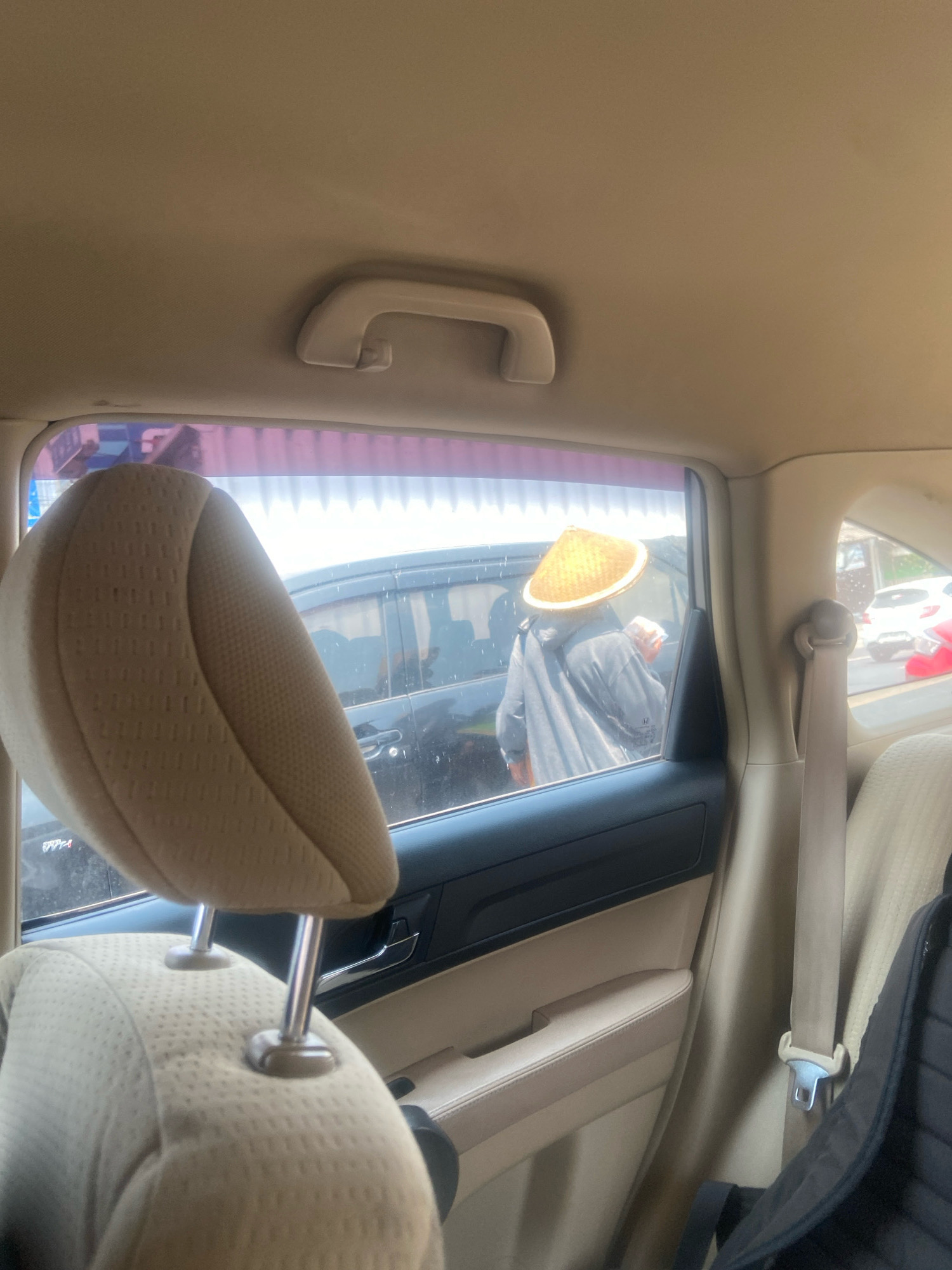 The image, taken from inside a vehicle, shows a person wearing a conical hat standing outside a black car. The person is wearing a gray hooded jacket and appears to be holding something in their hands. The view is through the car's side window, which has a tinted strip at the top. In the background, other vehicles are visible, including a white car and a red car, suggesting a street scene, possibly in the city of Ngaglik, Indonesia. The interior of the car is visible, with a beige headrest and part of a beige seat in the foreground. A white grab handle is attached to the car's ceiling. A black backpack is partially visible on the right side of the image, secured by a beige seatbelt. The overall lighting suggests it is daytime.
