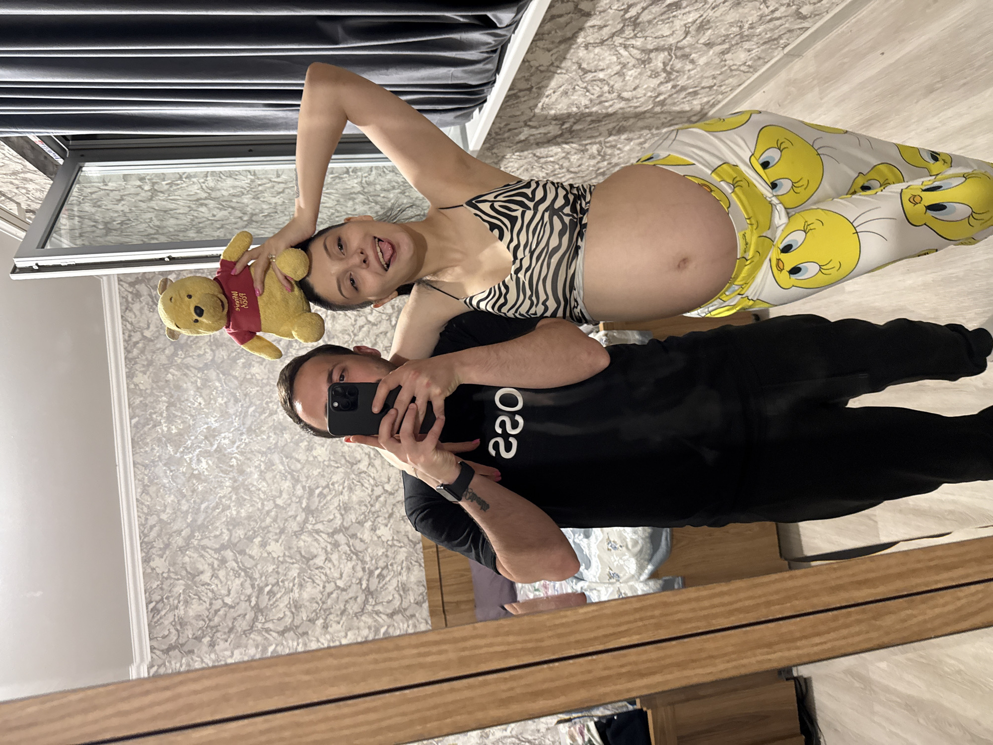 A man and a pregnant woman are taking a mirror selfie in a room that appears to be a bedroom. The man is on the left, wearing a black t-shirt and black pants. He is holding a smartphone to take the picture. The woman is on the right, wearing a zebra-print crop top and white pants with Tweety Bird graphics. She has a Winnie the Pooh stuffed animal balanced on her head and is sticking her tongue out at the camera. The room has wallpaper with a textured pattern, a wooden floor, a bed visible in the background, and a large window with grey curtains. The image was likely taken in Tbilisi, Georgia, given the prompt.