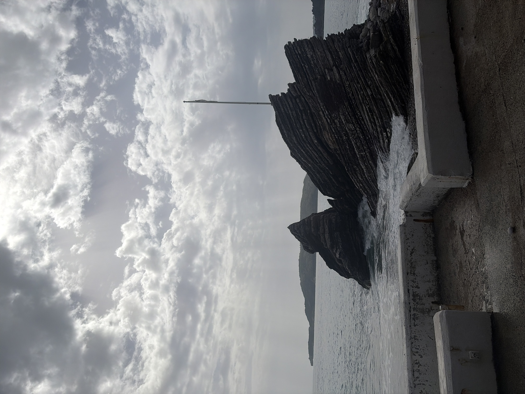 A coastal scene shows a rugged, dark rock formation situated in the water. Waves are crashing against the rocks and the base of a man-made structure. A pole extends upwards from the rock, reaching into the overcast sky filled with clouds. A concrete wall and part of a structure are visible on the right side. The location is in the city of Unknown, Unknown.