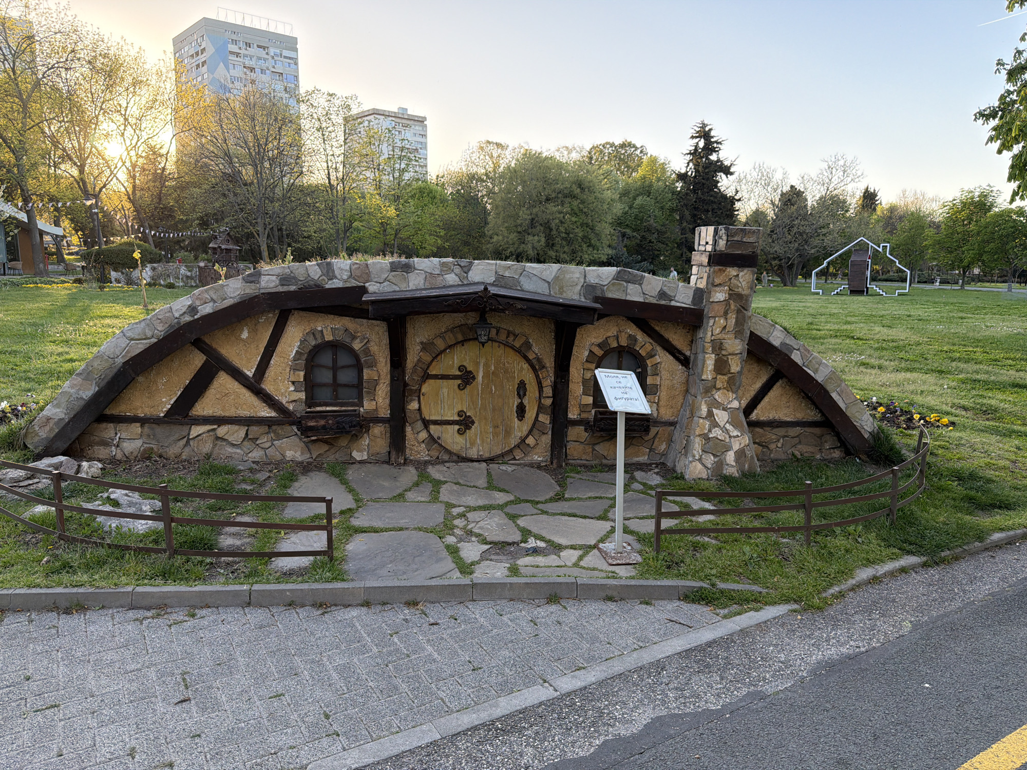 Captured at an eye-level perspective in the city of Unknown, Unknown, the image showcases a whimsical hobbit-hole-like structure. The facade features a round wooden door centered between two round windows, all set within a facade of stone and timber. A stone chimney is situated on the right side of the structure. A sign stands in front of the structure, and a low, curved metal fence surrounds the perimeter. The area directly in front of the hobbit-hole is paved with irregular stone slabs, interspersed with patches of grass. The structure is located on a grassy field within a park setting. In the background, modern apartment buildings rise above the trees, contrasting with the fairytale-like structure in the foreground. To the right of the structure, a white, outline-style sculpture of a house is visible in the distance. The scene is bathed in soft, natural daylight. A paved road runs along the bottom of the frame, indicating a public park or garden setting.