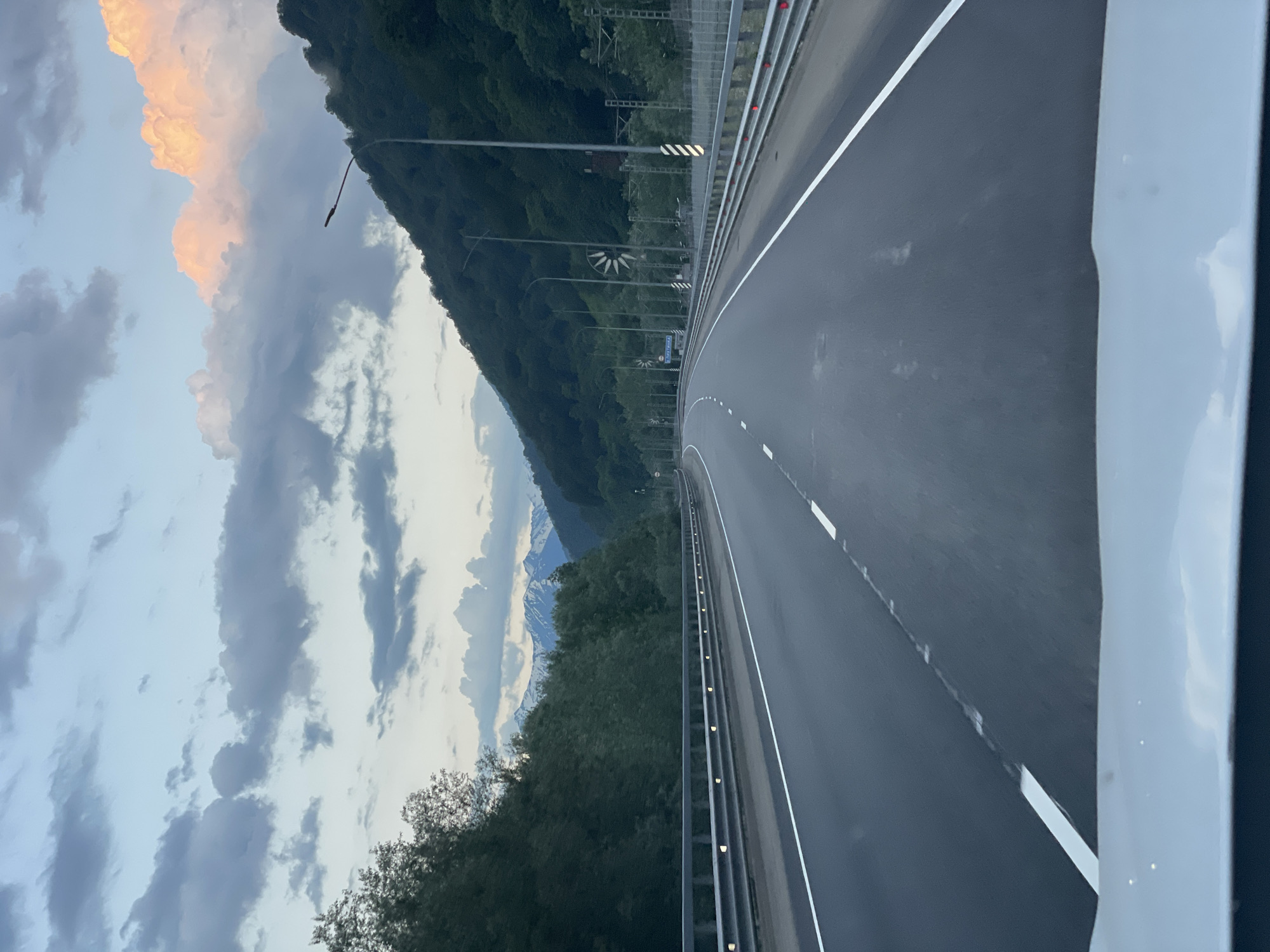 The image captures a view from inside a vehicle driving on a highway in Chvizhepse, Russia. The highway, with its lanes marked by white lines, curves to the right and is bordered by metal guardrails. On the left, a dense forest of trees lines the road, and on the right, a steep, forested mountainside rises dramatically. In the distance, snow-capped mountains are visible against a sky filled with dramatic clouds, some tinged with pink from the setting sun. Several lampposts line the highway, some with what appear to be wind-powered turbines attached. The overall scene suggests a scenic drive through a mountainous region during twilight.