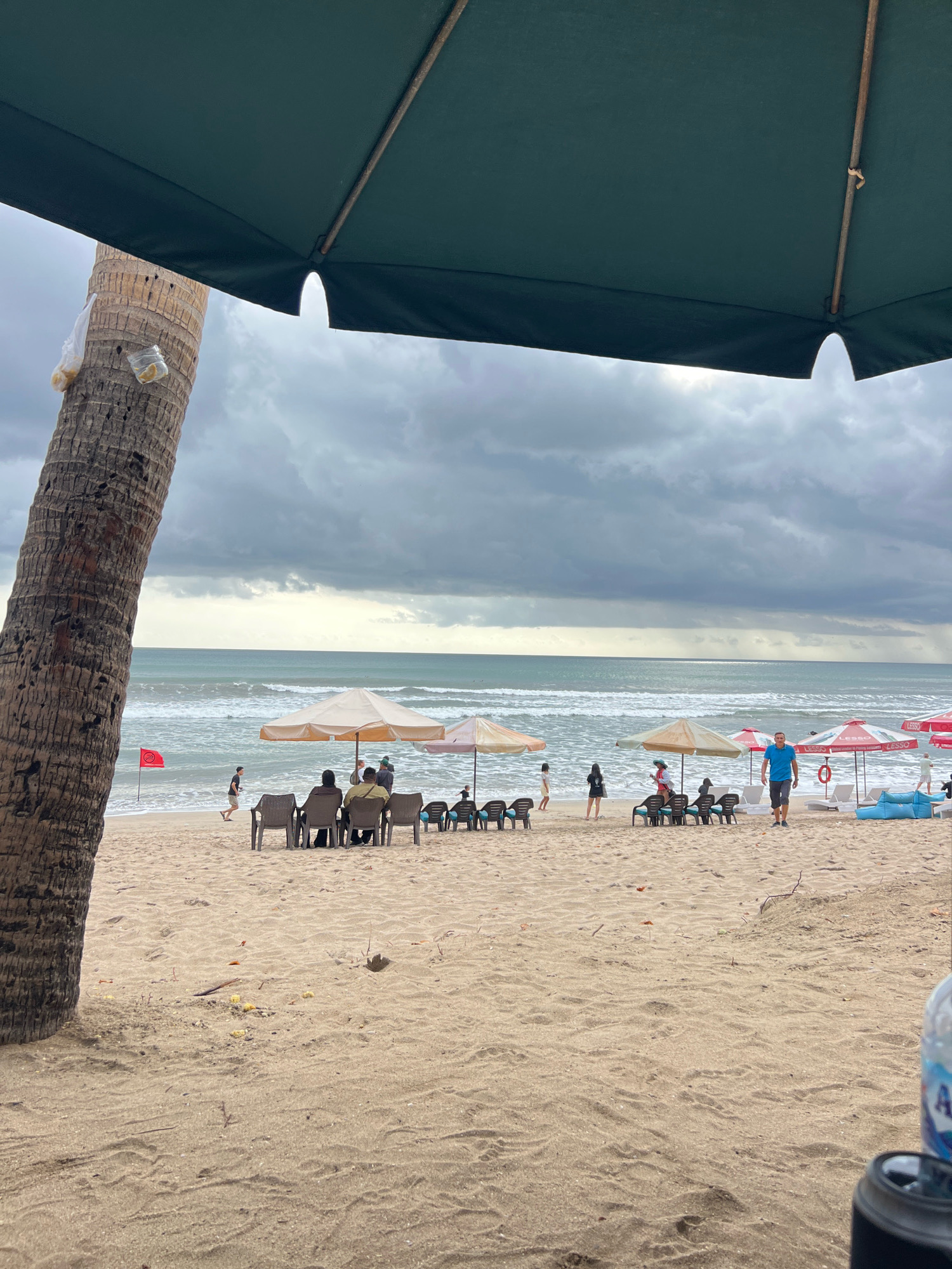 An eye-level medium shot captures a beach scene in Kuta, Indonesia, under a cloudy sky. The foreground is dominated by the textured sand of the beach, with a large portion of a dark green umbrella and a thick palm tree trunk on the left, partially obscuring the view. The umbrella's brown metal pole is visible. On the palm tree, two plastic bags are tied, one with a few pieces of food inside.
The middle ground reveals a stretch of sandy beach with several beach umbrellas providing shade. Under one of these umbrellas, a group of people are seated in chairs, facing the ocean. In the distance, the ocean has moderate waves, and several figures are visible on the sand, including a man in a blue t-shirt and dark shorts walking towards the viewer, a person in a red life preserver, and other individuals scattered on the beach, some sitting or standing under umbrellas. A red flag is planted in the sand on the left side of the image.
In the background, the ocean extends to the horizon, meeting the overcast sky. The lighting is diffused due to the clouds, creating a subdued atmosphere. The overall scene depicts a relaxed beach environment with people enjoying the coastal scenery.