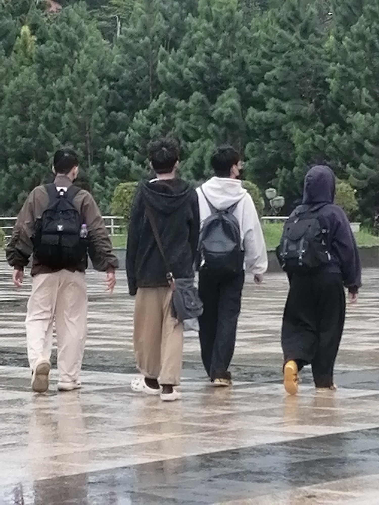 Four individuals walk away from the camera on a wet, paved surface. The individuals are dressed casually and each carries a backpack. The background consists of dense green trees. Based on the architectural style of the paving and the presence of dense evergreen trees, this scene appears to be located in Da Lat, Vietnam, possibly at a park or a promenade. The person on the far left wears a brown jacket and beige pants. The second person from the left wears a black hoodie and beige pants, with a crossbody bag. The third person from the left wears a light-colored hoodie and black pants. The person on the far right wears a dark hooded jacket and black pants. The ground is reflective, indicating recent rain.