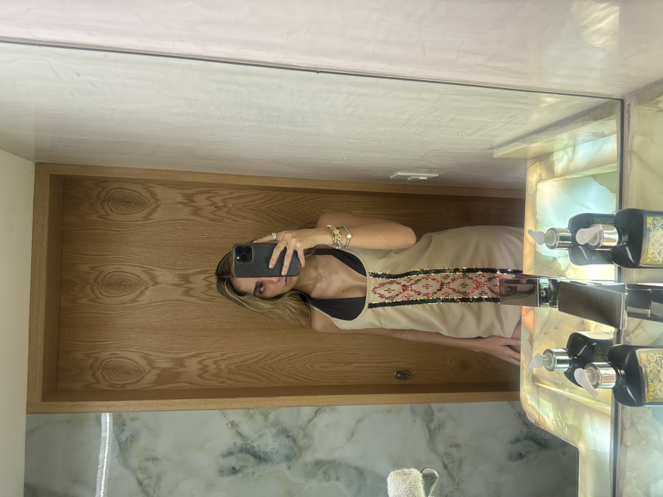A woman takes a mirror selfie in a bathroom. She holds a phone, her face visible behind it. She wears a tan dress with a black top, featuring an embroidered horizontal pattern with red and black details on the bodice. The bathroom features a light wood vanity, marble countertop, and a prominent soap dispenser. The reflection shows the bathroom's interior, including towels and other fixtures.