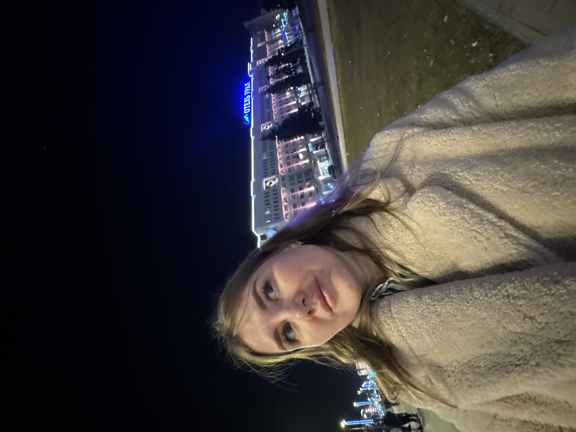 A woman wearing a light beige coat is posing for a selfie at night in Perm, Russia. In the background, the illuminated "Hotel Ural" building is visible. The sky is dark, and there are lights along the embankment. The woman is looking at the camera.