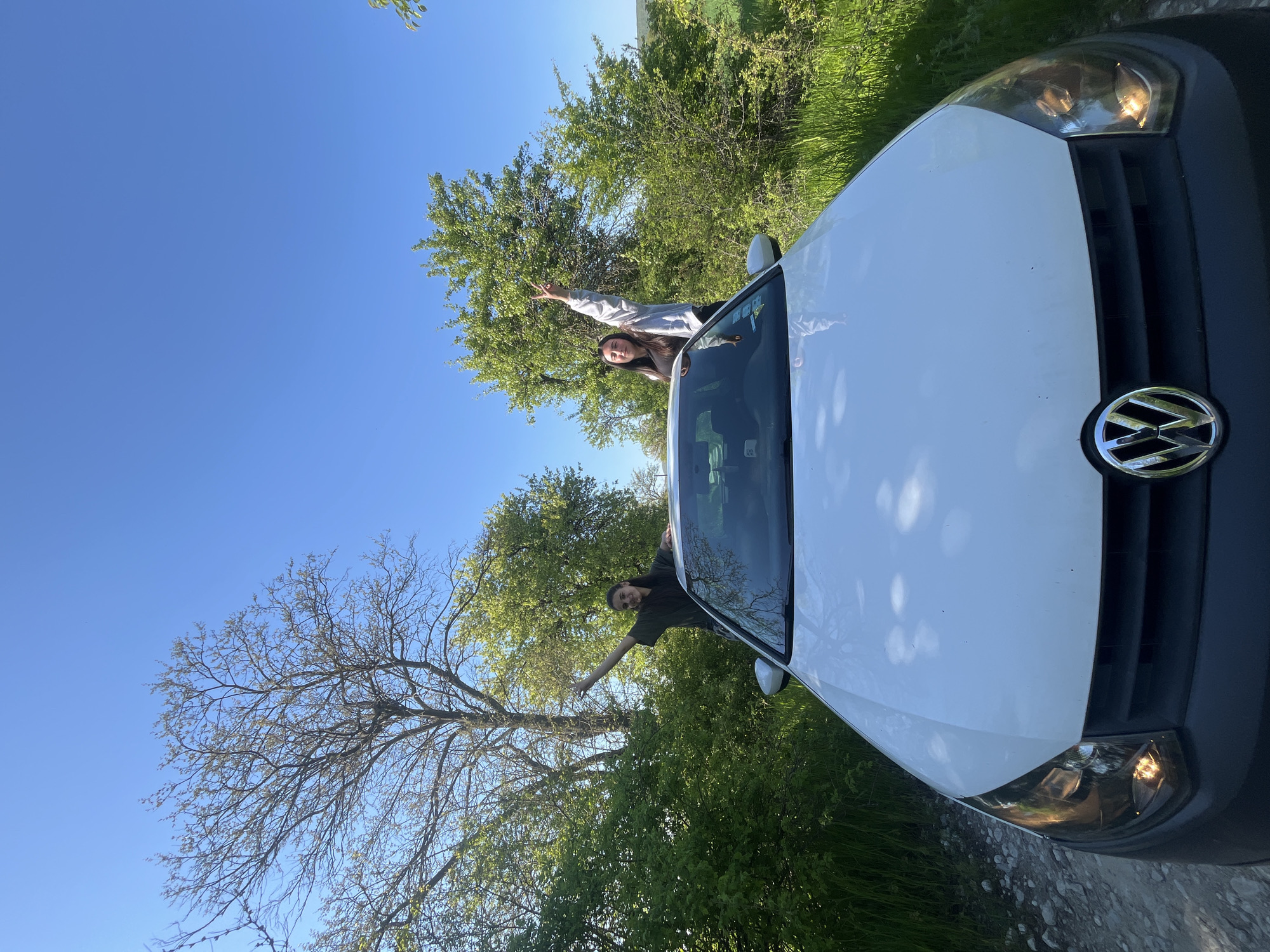 Two people are leaning out of the sunroof of a white Volkswagen automobile. The vehicle is parked on a paved road next to green grass, bushes, and trees under a clear blue sky. Based on the vehicle make, model, and scenery, the location is likely Rish, Bulgaria.