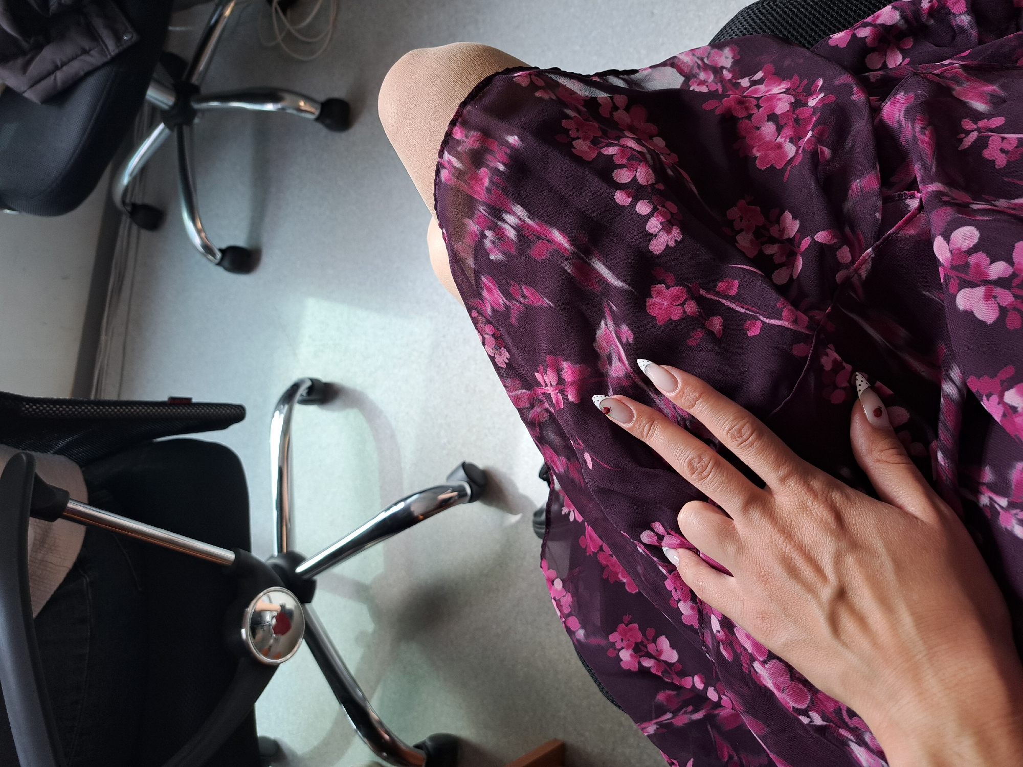 This high-angle shot captures a person seated in an indoor setting, likely an office or similar workspace in Sofia, Bulgaria. The central focus is the person's lap, adorned with a dark purple dress or skirt featuring a prominent pink floral pattern. One leg, possibly covered in light-colored hosiery, is visible above the garment. A hand with manicured nails, styled with a French tip and some accented with small red heart details, rests gently on the fabric.
Two office chairs with black mesh or padded seats and five-spoke chrome bases with black wheels are visible. One chair is partially in the foreground on the left, while another occupies the upper left corner, with a dark jacket or coat draped over its backrest. The floor beneath the chairs is a light grey or beige, possibly concrete or linoleum, showing shadows that suggest natural light entering the room. The image conveys a calm, everyday scene in a functional environment. No discernible text is present.