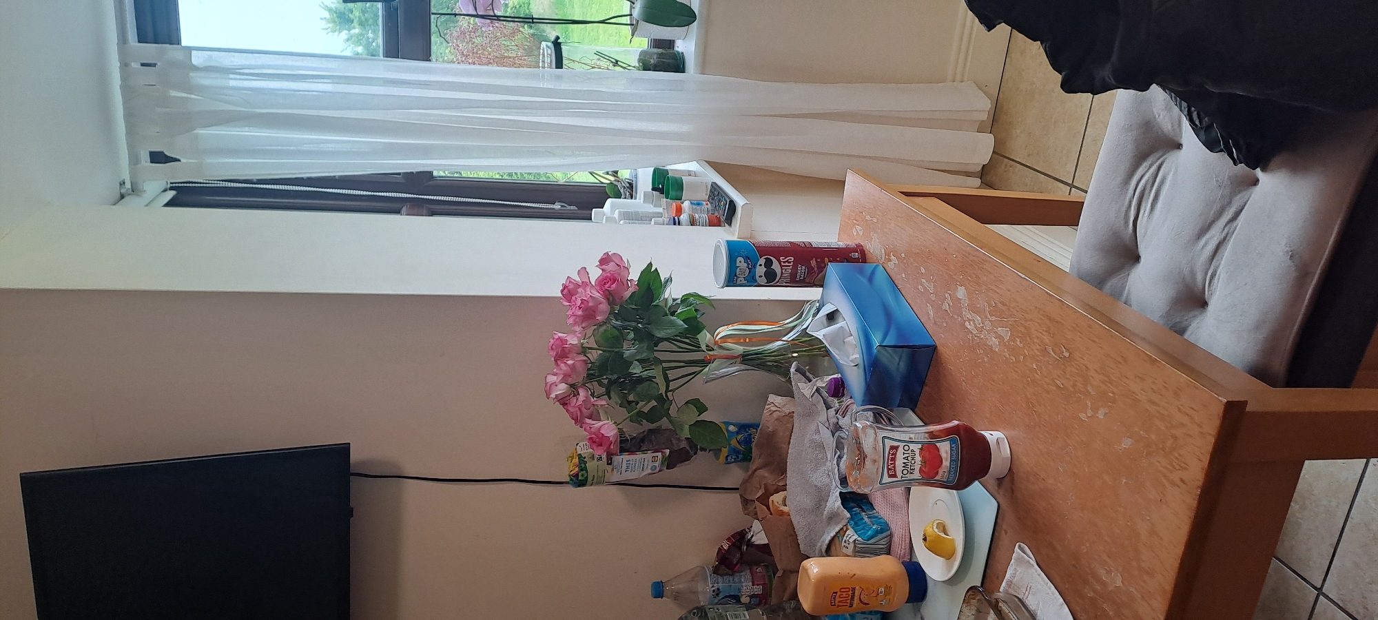 The image depicts a domestic scene, likely a kitchen or dining area. A wooden table occupies the foreground, with a padded chair positioned underneath. On the table, various items are arranged, including a bottle of tomato ketchup, a lemon wedge on a plate, a bottle of mayonnaise, a box of tissues, and a tube of Pringles. A bouquet of pink roses in a vase adds a touch of color. In the background, a window with white curtains offers a view of greenery. A television is mounted on the wall to the left. The overall impression is one of casual clutter, suggesting recent use. The location is identified as Castlebar, Ireland.