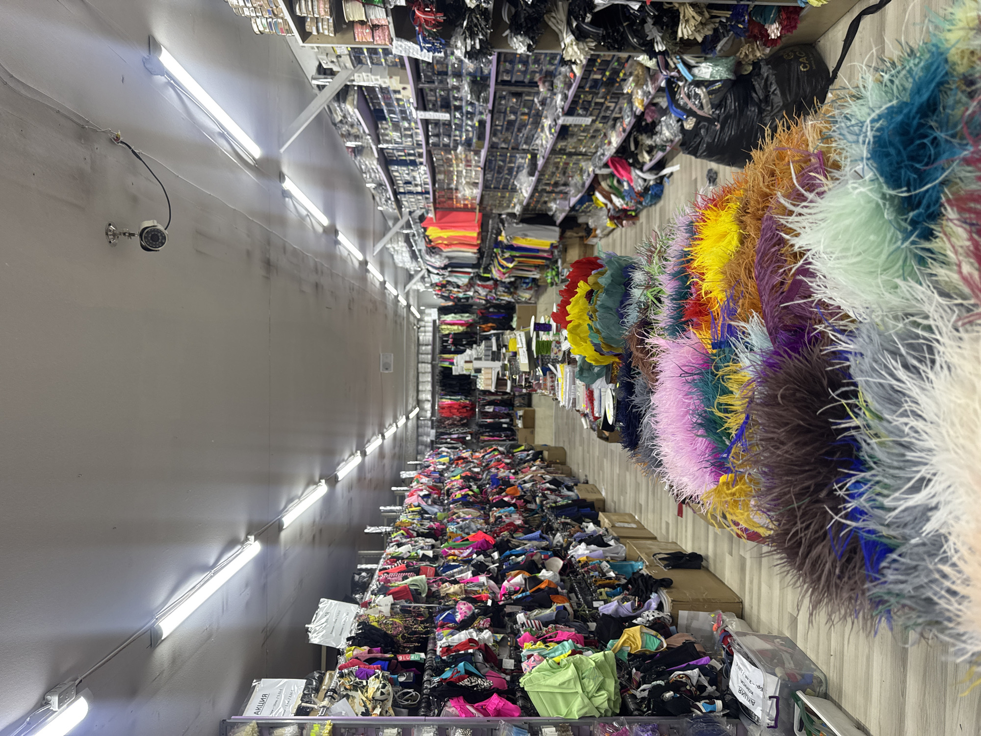 The image shows the interior of a narrow warehouse or shop in what appears to be Sofia, Bulgaria, stocked with a variety of craft and sewing materials. The scene is brightly lit by a series of fluorescent tube lights running along the ceiling. Security camera is installed on the left wall.
Shelves on either side of the narrow aisle are densely packed. On the left are rows of small items packaged in individual containers, likely beads, buttons, or other embellishments. The bottom shelves are cluttered with bundles of fabric scraps and trim.
On the right, colorful ostrich feathers of various hues are displayed. These appear to be the focal point of the shop's inventory. A black garbage bag and other storage items are also visible on the right.
The aisle floor is also cluttered, adding to the sense of a busy, perhaps somewhat disorganized, retail or wholesale space. Some boxes are visible on the floor.
The overall impression is of a specialized shop catering to the crafting or fashion industries, with a concentration on trimmings and decorative materials. The Russian language text on the packaging suggests the origin of the item.