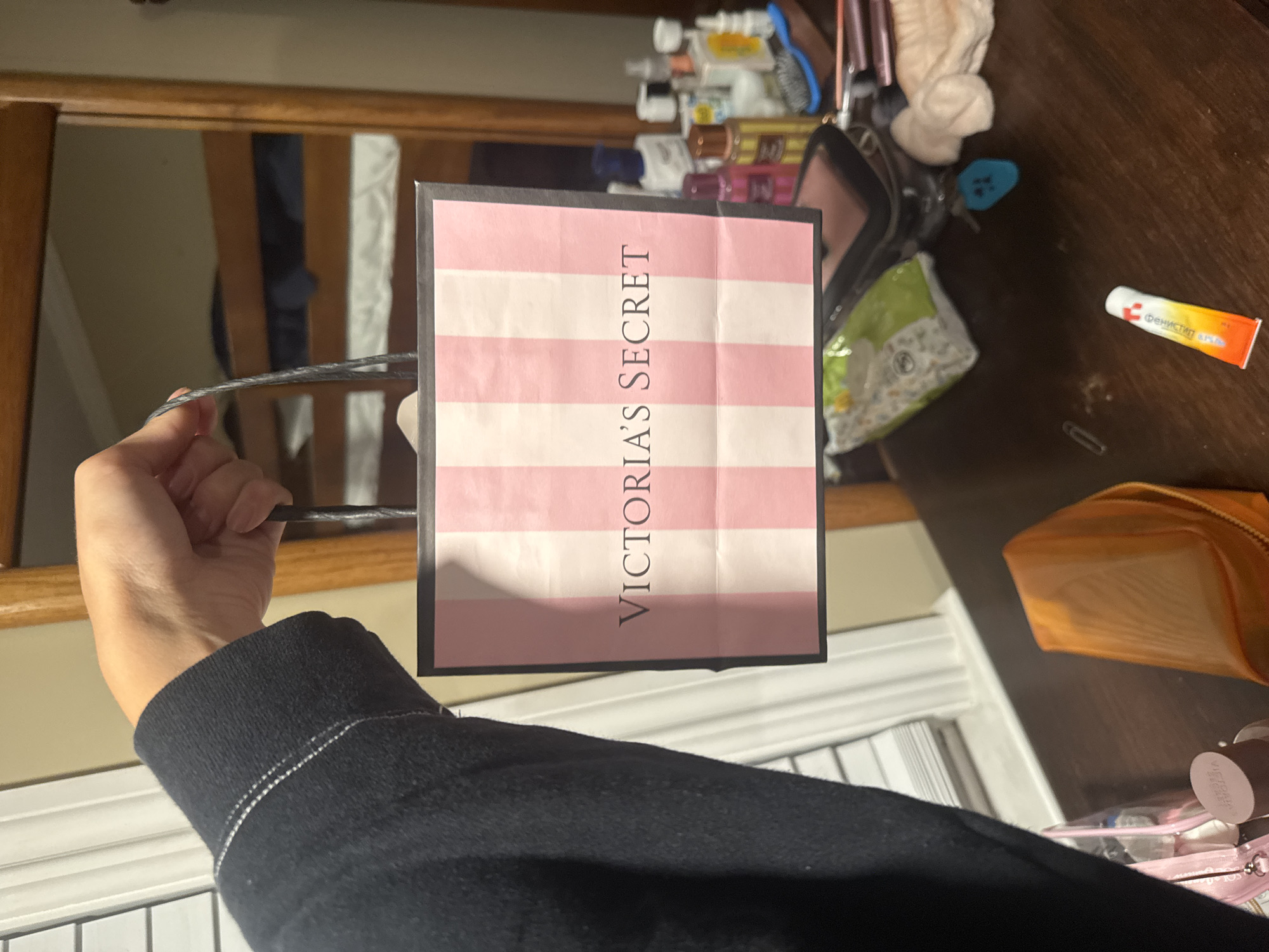 A person's hand and arm, wearing a black hoodie, holds up a small pink and white striped shopping bag with "VICTORIA'S SECRET" printed on it. The bag is the primary subject in the foreground. The background reveals a blurred indoor environment, possibly a bedroom or dorm room, with bunk beds visible. Various objects are scattered on a wooden surface below, including cosmetic bottles, a makeup bag, and what appears to be a tube of ointment. The overall lighting suggests an indoor setting at night or in low light conditions. No specific location in Ocean City, United States of America is depicted.