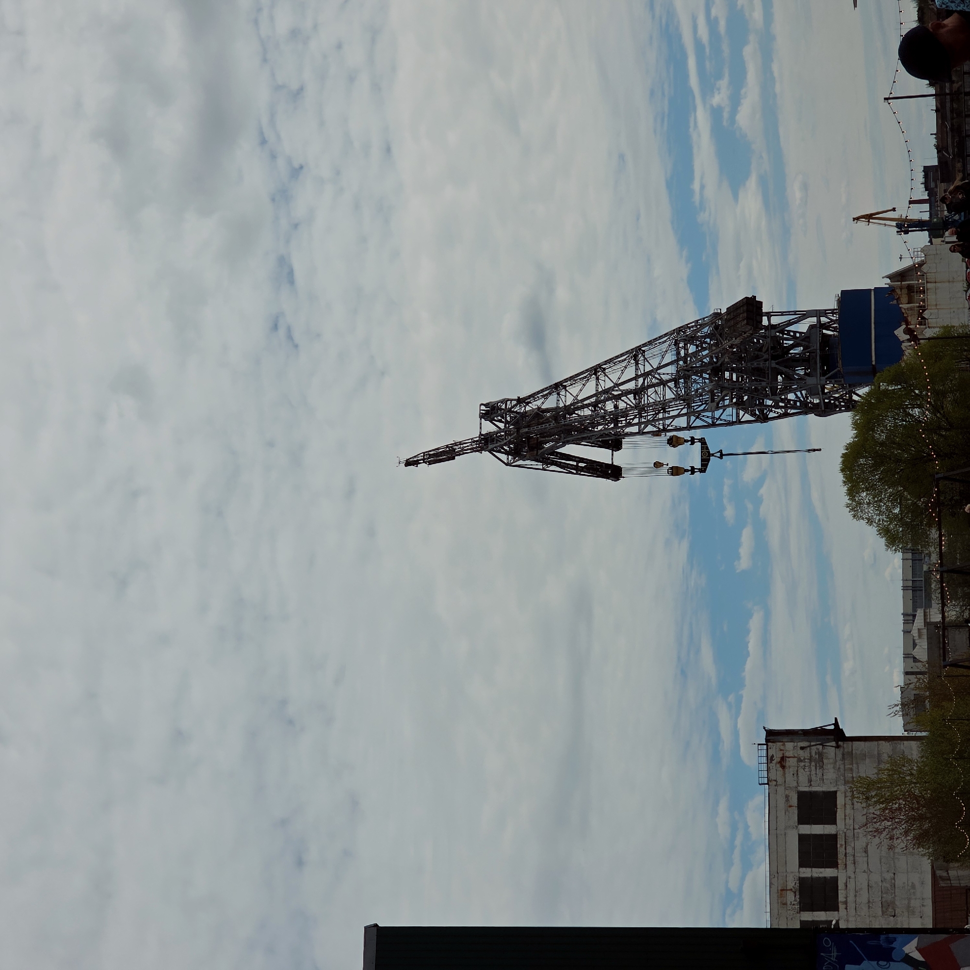 The image shows a large crane set against a cloudy sky, likely situated in an industrial or port area. The crane is made of a complex network of steel beams, and its boom extends outward. In the background, there are buildings, including one with several windows and weathered facade, and other cranes or structures. There are trees in the foreground. The scene depicts an environment typical of a working harbor or dockyard. While the image does not provide explicit confirmation, it resembles a location along the Neva River, within the city of Saint Petersburg, Russia.