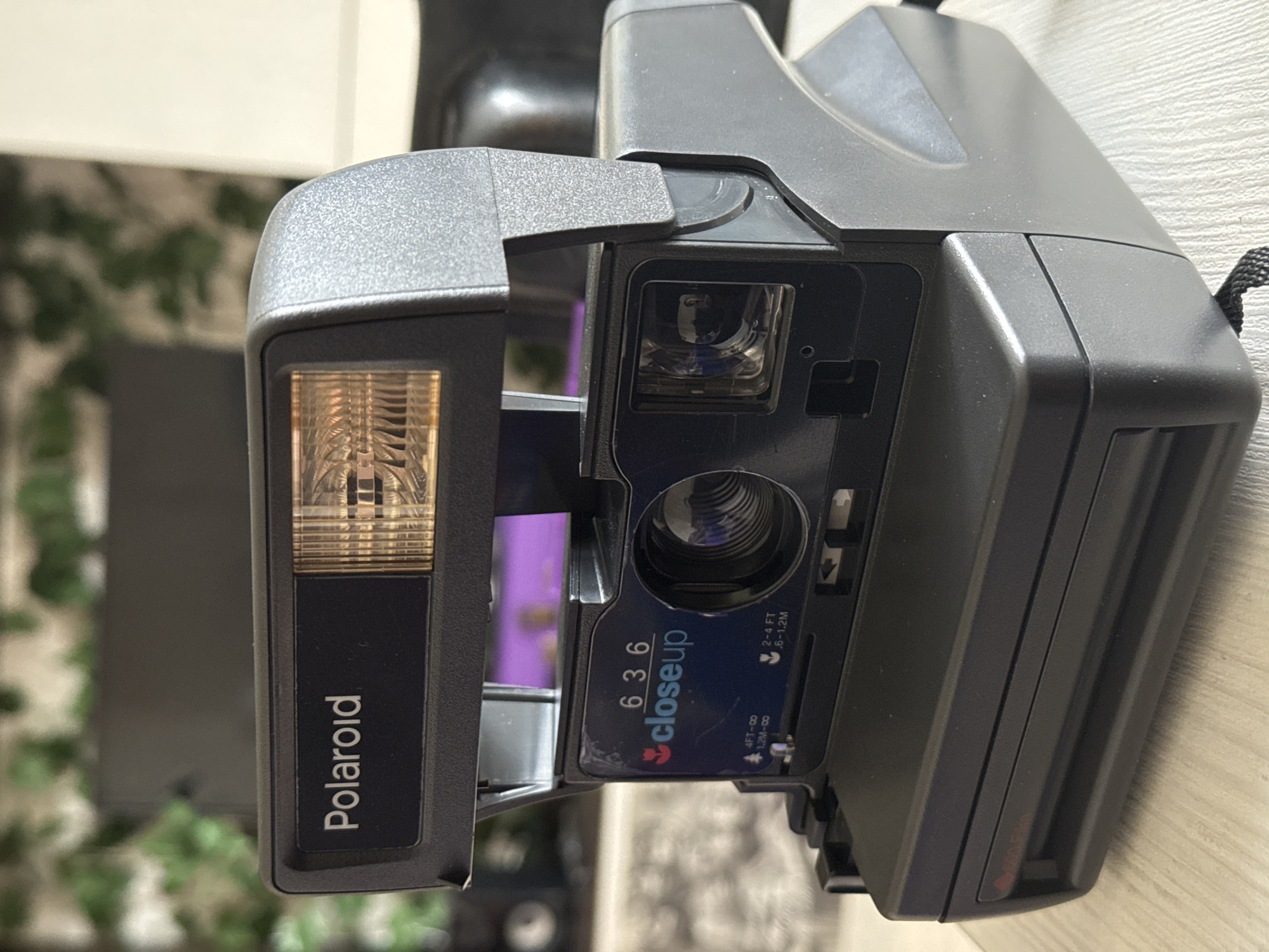 The image shows a Polaroid 636 closeup camera. The camera is positioned on a light-colored, possibly wooden surface. The flash housing is visible on the left, with the "Polaroid" logo prominently displayed. The lens area has a blue sticker with "636 closeup" printed on it, along with distance measurements in feet and meters. In the background, there's greenery suggestive of an indoor plant or outdoor environment visible through a window. There are no people in the frame. The location of Novo-Nikol'skoe, Russia, is not discernible from the image.