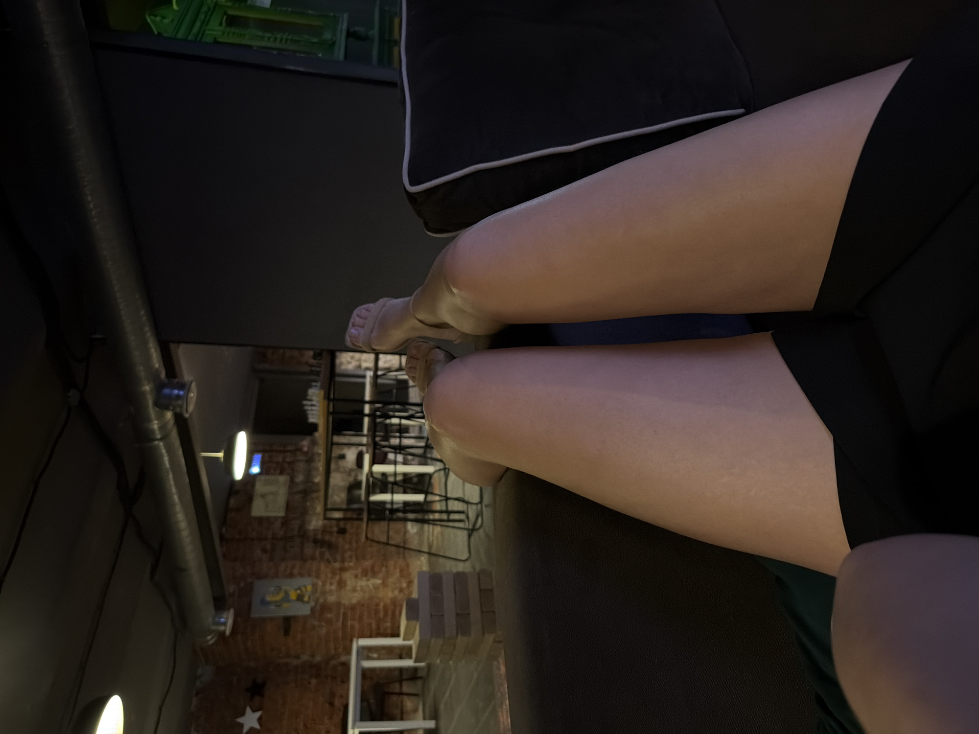 A low-angle, horizontally oriented photograph depicts a partial view of a person seated on a dark-colored sofa. The subject's legs, clad in what appears to be dark shorts, are prominently featured. The background includes a red brick wall, various shelves, and an industrial-style ceiling with pipes and lights. The specific location within Moscow, Russia cannot be determined from the image alone.