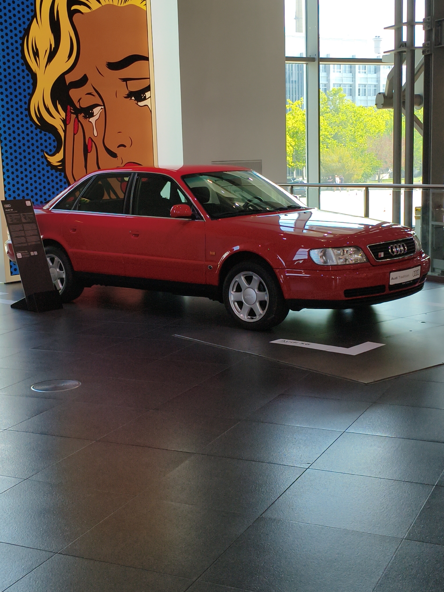 A red Audi S6 sedan is on display in a brightly lit modern building with large windows. The car is positioned on a dark tiled floor, which reflects the light. Behind the car, a large pop art-style painting depicts a crying woman with blonde hair and yellow skin. To the left of the painting, a black display stand with information about the car is visible. Through the windows, trees with bright green foliage and modern buildings can be seen. The overall impression is that of a car museum or exhibition space. The building appears to be part of the Audi Forum Ingolstadt, a visitor center for Audi, located in Ingolstadt, Germany. No people are clearly visible interacting with the car or the artwork.