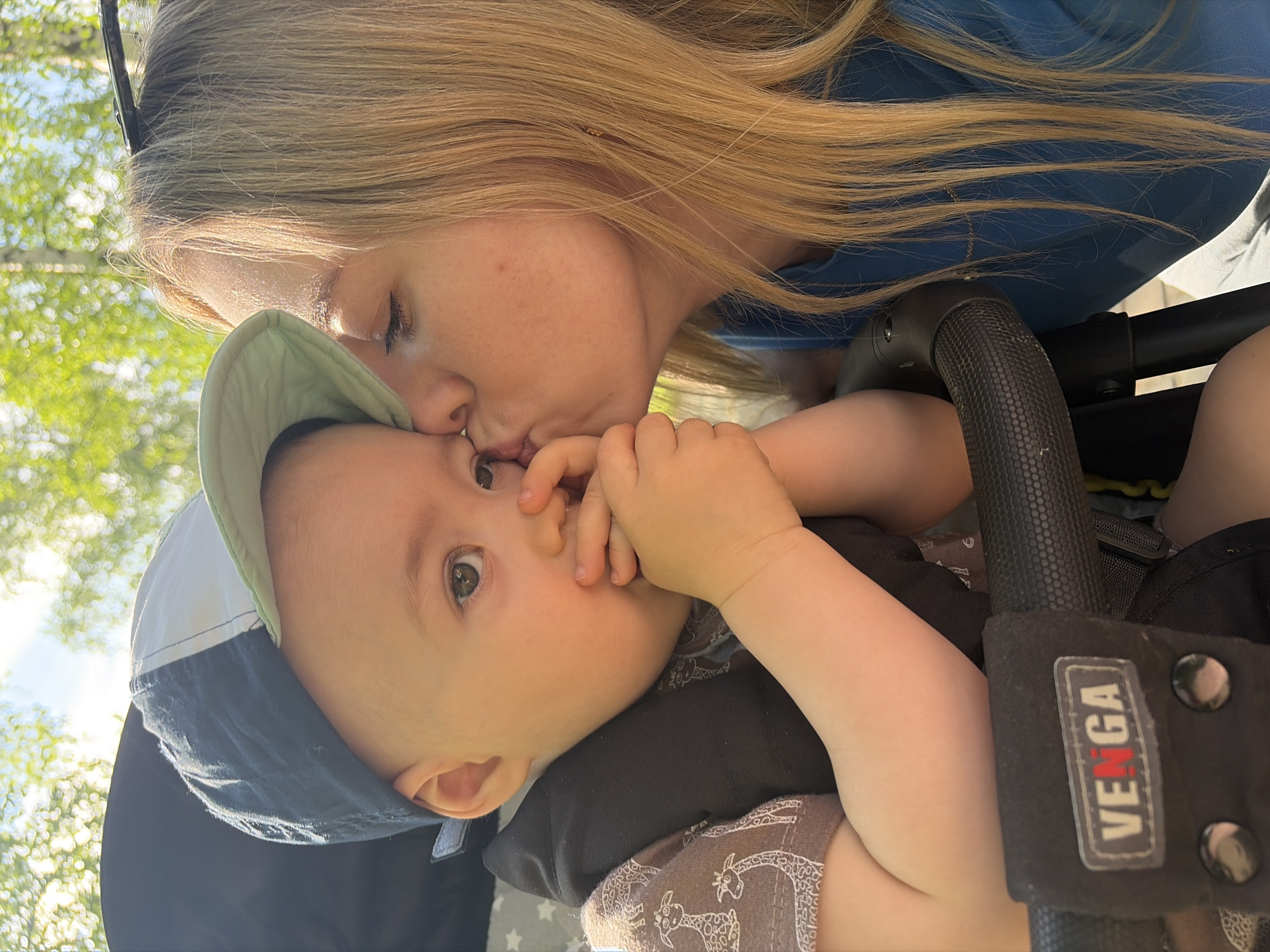 A woman is kissing a baby in a stroller. The woman has blond hair and is wearing a blue shirt. The baby is wearing a hat and has their fingers in their mouth. The stroller has a "VENGA" logo on the side. The location appears to be outdoors, possibly in a park, with trees visible in the background. It is unclear whether this is the city of Chaikovsky, Russia, as there are no definitive landmarks.