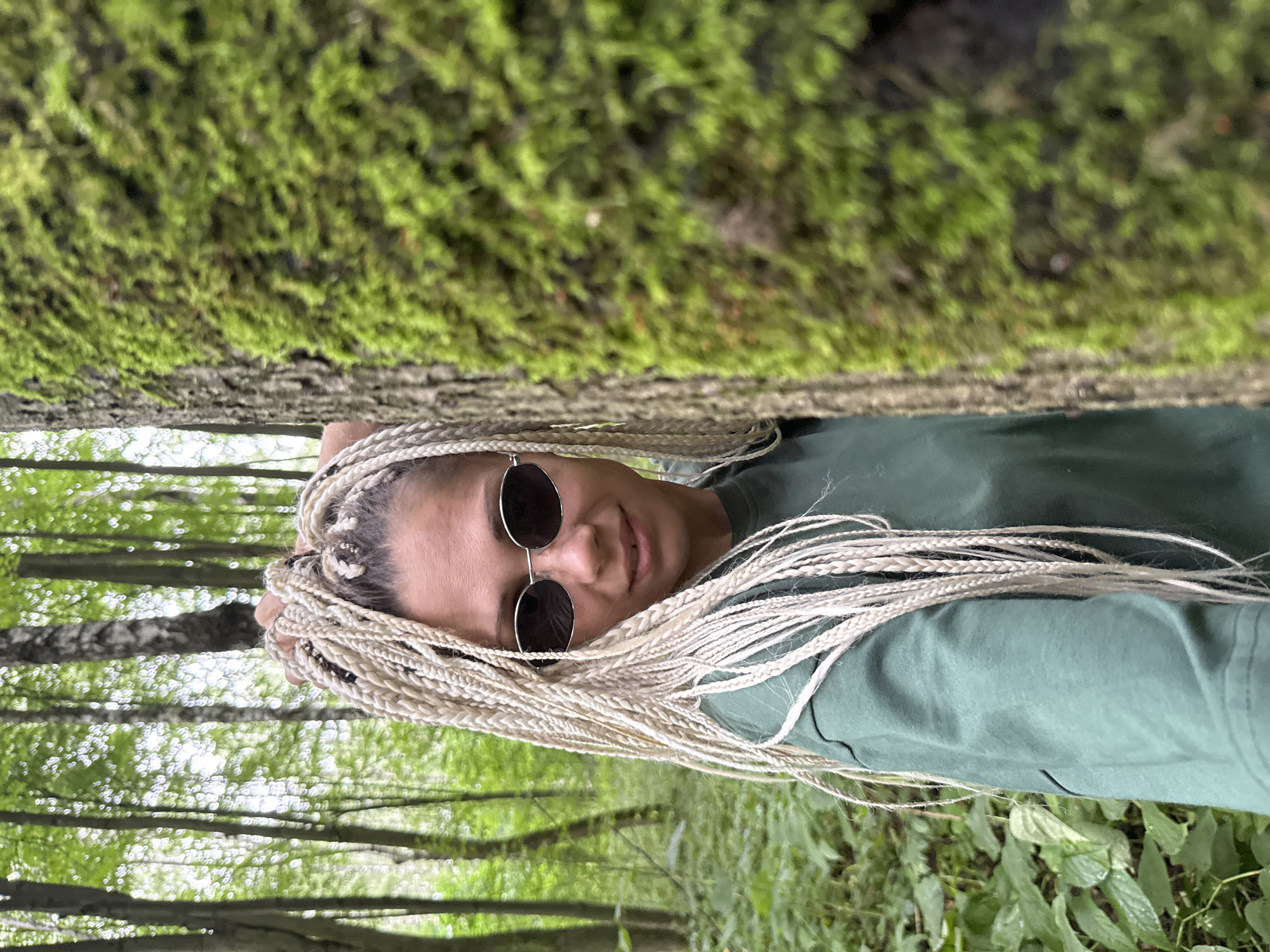 A woman with blonde braided hair and sunglasses is partially obscured by a moss-covered tree in a forest. She is wearing a green sweatshirt and looking towards the camera. The background consists of tall trees and green foliage, typical of a forest environment. The specific location is Rusanovo, Russia.