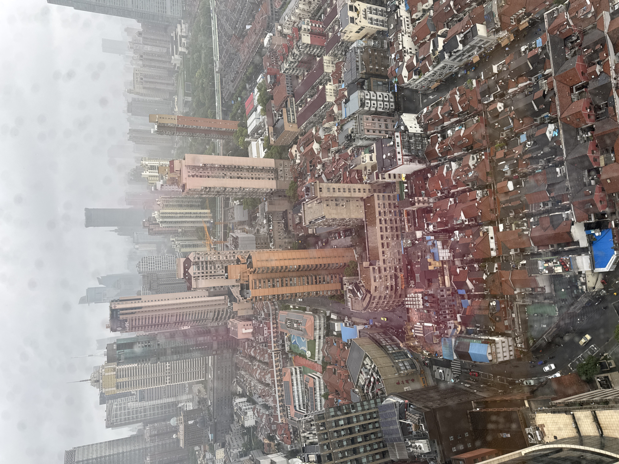 This is an aerial view of a densely populated urban area in Shanghai, China, likely taken on a rainy or overcast day, as indicated by water droplets on the lens and the muted light. The cityscape is a mix of old and new architecture. Tall, modern skyscrapers with glass facades dominate the background, interspersed with older, lower-rise buildings featuring red-tiled roofs. In the foreground and mid-ground, a dense cluster of residential buildings with red and dark grey tile roofs covers much of the landscape, with narrow streets winding between them. Several vehicles, including cars and what appear to be motorcycles, are visible on the wet streets. Some buildings have solar panels installed on their roofs, suggesting a move towards renewable energy. The overall impression is one of a bustling city with a long history and continuous development.