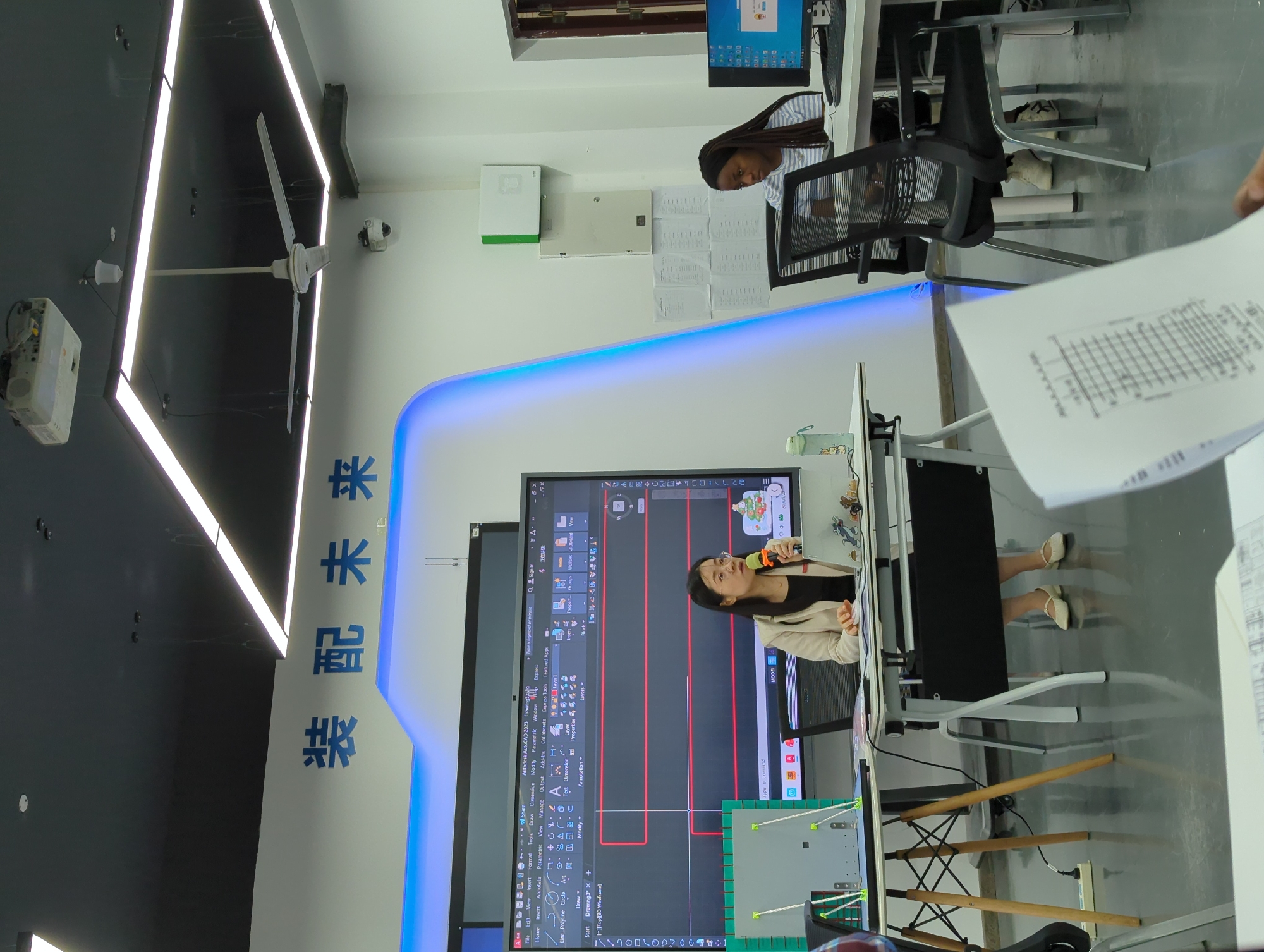 The image shows an office-like environment in Jiangjin, China. A woman is presenting in front of a large screen displaying AutoCAD software, using a microphone. There are two other people seated at desks with computers. The room features modern lighting fixtures, including ceiling lights and a blue-lit panel. The Chinese characters "装配未来" are prominently displayed on the wall. The scene appears to be a training session or a demonstration of CAD software. There are desks and office chairs. Someone is holding a graph document near the right side of the frame.