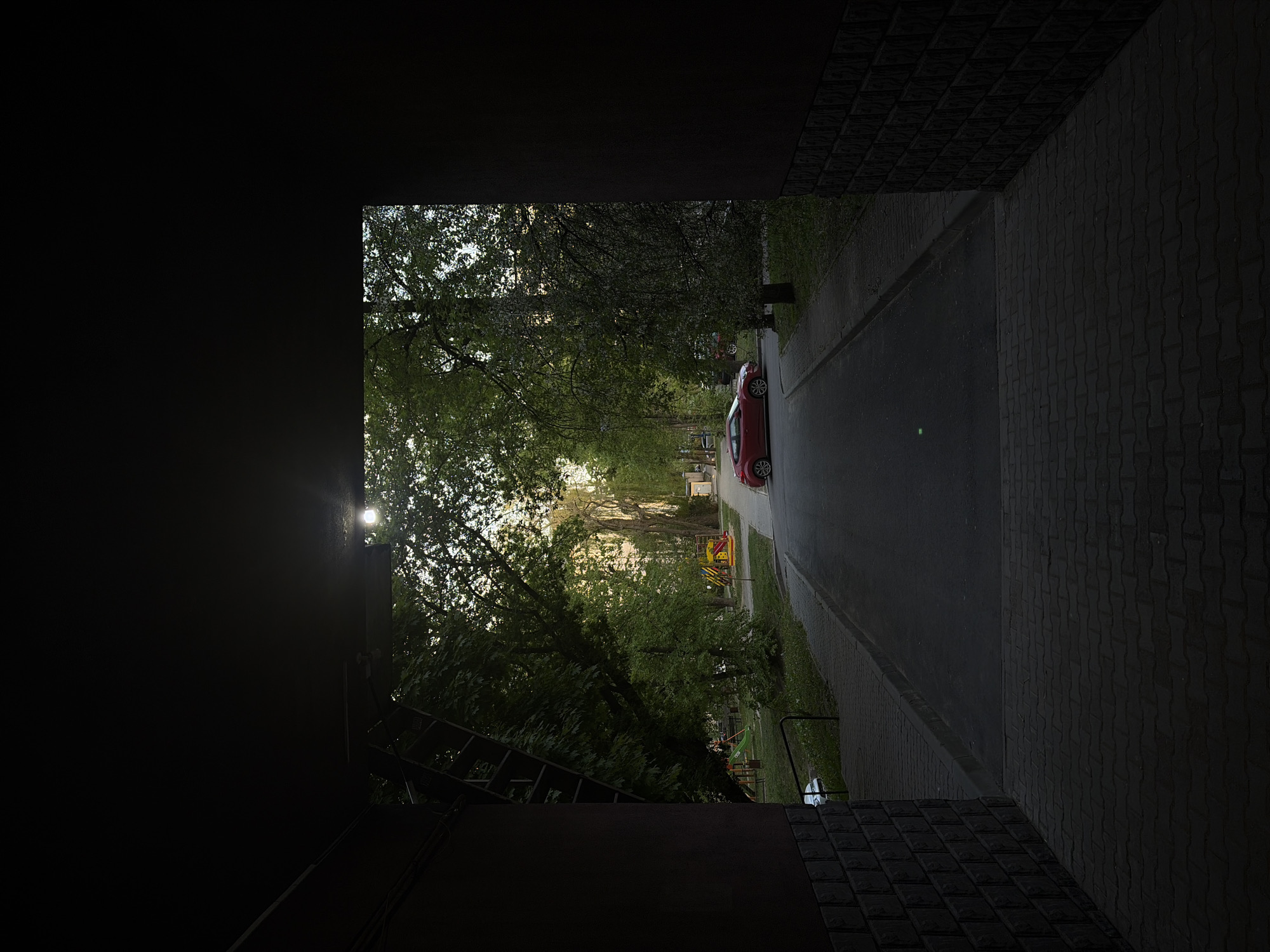 View from inside a dark structure, looking out onto a street lined with trees and a red car parked on the side. In the distance, a playground can be seen. The location is Unknown, Unknown.