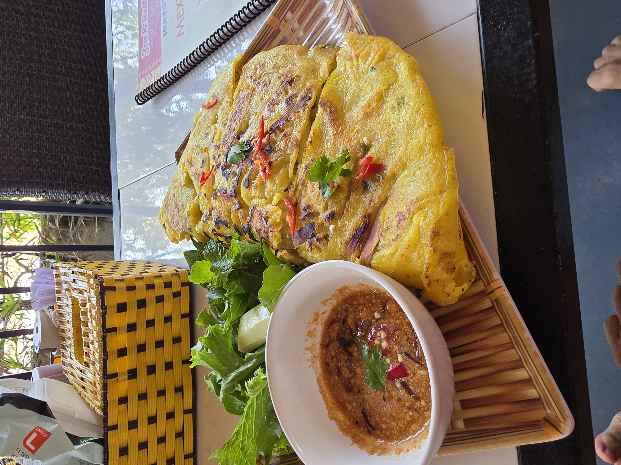 A high-angle, slightly wide-shot photograph shows a dish of Vietnamese banh xeo served on a woven, triangular platter on a table, likely at a restaurant. The dish is accompanied by a small bowl of dipping sauce and fresh herbs. Part of a menu is visible in the background, as well as a woven container. A sliver of a foot is seen on the right edge of the frame.
The banh xeo, a savory crepe or pancake made with rice flour, coconut milk, and turmeric, is folded in half and cut into sections. It is garnished with sprigs of fresh parsley and slivers of red chili.
The dipping sauce, served in a white bowl, is a light brown color and speckled with chili pieces and a sprig of fresh parsley.
The herbs, a vibrant green color, surround the dipping sauce and part of the banh xeo. They appear to be a mix of lettuce and mint.
In the background, to the left of the plate, is a yellow woven container with a black grid pattern. Further back, part of a menu or sign is visible, along with the top of a chair. The table has a white, reflective surface.
The setting suggests an outdoor dining area in Mũi Né, Vietnam, likely at a restaurant specializing in local cuisine.