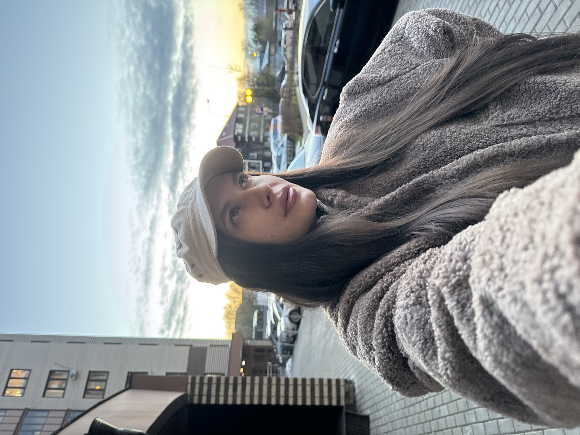 The image, rotated 90 degrees to the left, captures a young woman in an urban setting, likely Odintsovo, Russia, taking a low-angle selfie. She is positioned on the right side of the frame, looking upwards and to her left (our right).
The woman is wearing a light beige baseball cap and a thick, textured, fluffy jacket in a warm gray or brown color. Her long, dark brown hair cascades over her shoulder and arm. Her face is visible, featuring clear skin and full lips, with her gaze directed towards something in the upper distance.
The background consists of a clear blue sky with scattered wispy clouds in the upper left section, suggesting a bright day. The lower right section reveals an urban environment with buildings, some featuring illuminated windows. Cars are parked along a street or parking area. A warm, golden light is visible in the distant background, indicating either sunrise or sunset, with the sun positioned low on the horizon. Streetlights are also discernible. The foreground shows paved ground with a textured, brick-like pattern. A faint "P" sign, likely for parking, is visible in the distant background. The scene conveys a calm, early morning or late afternoon atmosphere in the city.