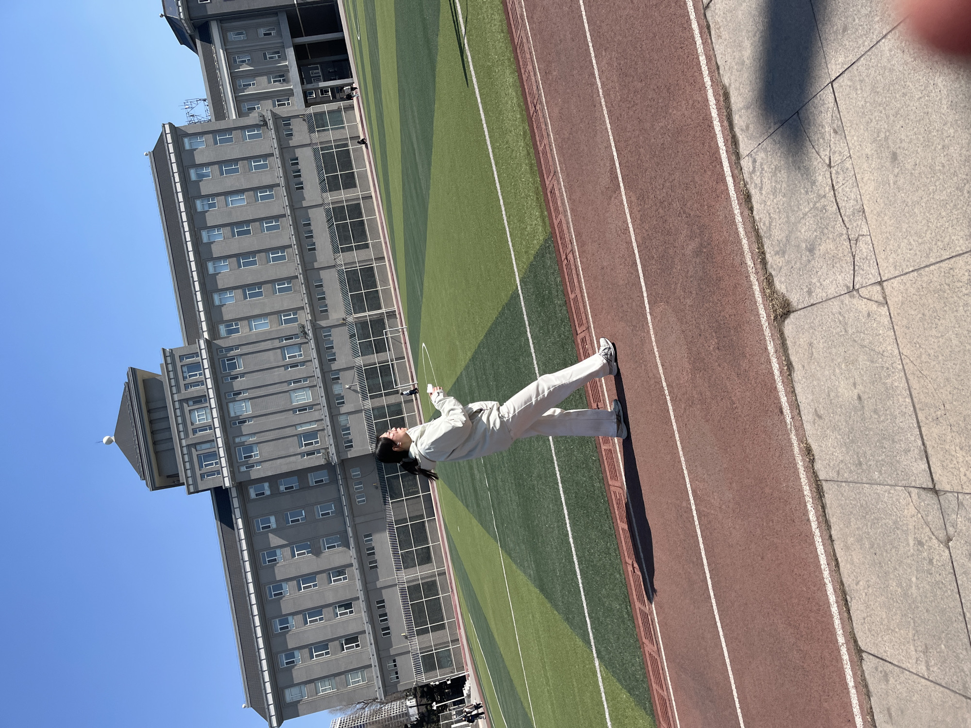 A young woman stands on the red running track adjacent to a green sports field. She is dressed in light-colored clothing and appears to be looking up. The background features a large gray building with multiple windows, indicating an educational or institutional structure. Above the building is a clear blue sky. The scene is located in Changchun City, China, identifiable by the architectural style and overall urban landscape.
