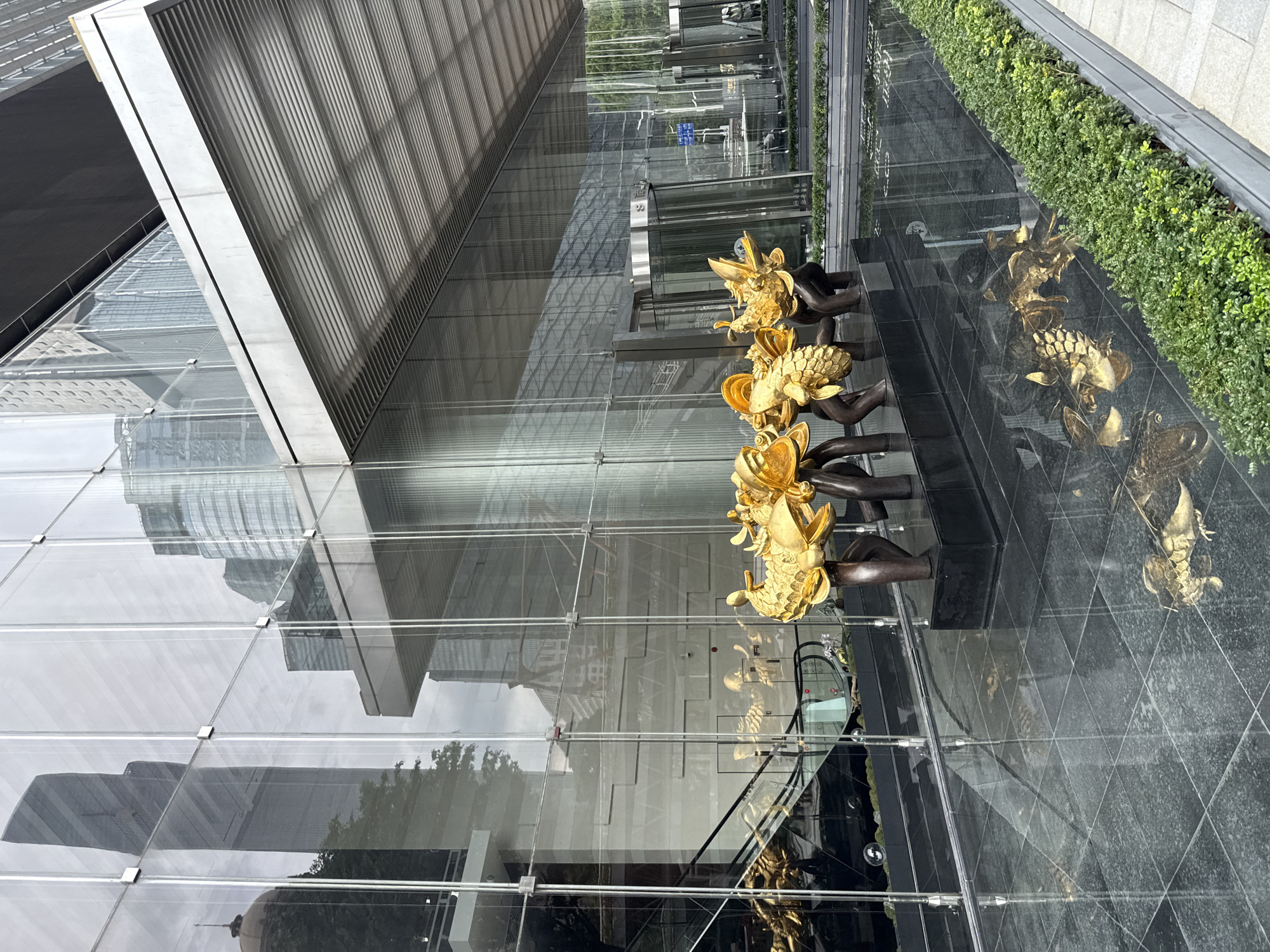 This image captures the exterior of a modern office building in Tianhe District, China. The building features a facade of large glass panels that reflect the surrounding urban landscape, including other skyscrapers and trees. In front of the building, a water feature with dark, polished stone surfaces holds several gold-colored sculptures, seemingly of mythical creatures like dragons or griffins, positioned on a dark base. The sculptures are reflected in the water. Adjacent to the water feature is a neatly manicured green planter box filled with lush vegetation. An escalator is visible through the glass on the left side of the building. A revolving glass door serves as an entrance to the building on the right, and a street sign is visible through the glass. The overall impression is one of contemporary urban design and public art.