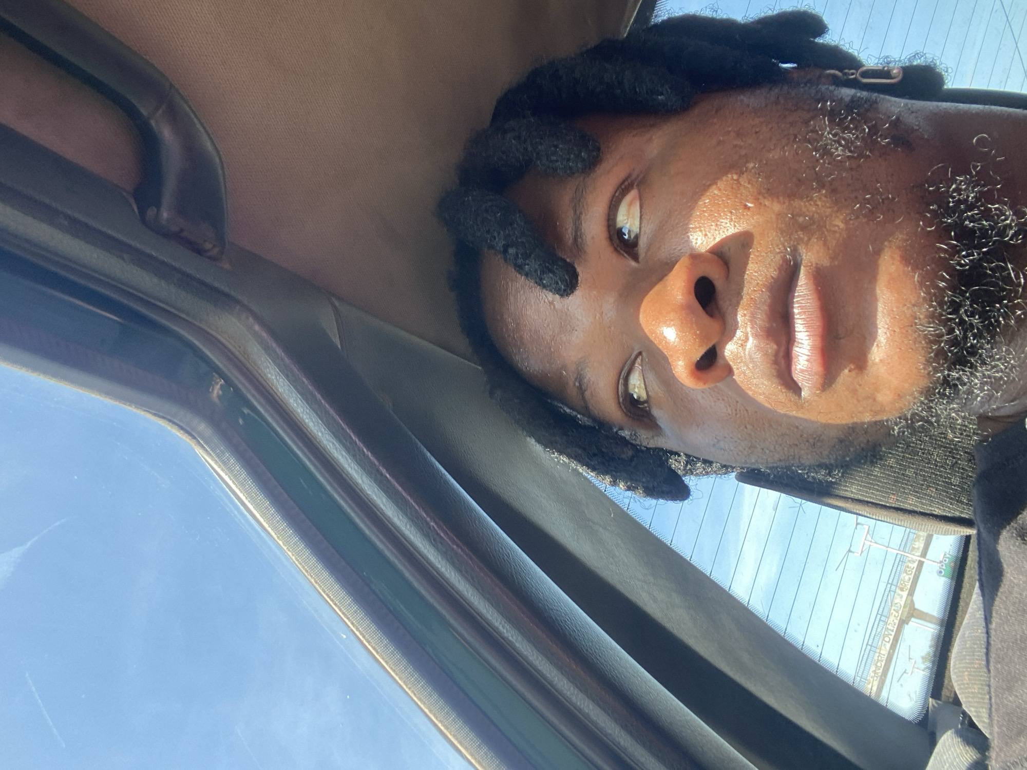 The image is a close-up, upward-angled shot of a Black person with dreadlocks, looking out of a vehicle's sunroof. The person's face is illuminated by sunlight, highlighting their skin texture and some facial hair. They are wearing a dark shirt, and a silver earring is visible on their right ear. The background shows a bright blue sky through the sunroof and the rear window. Reflected in the rear window, a road, an overhead bridge with writing, and some signage are visible. The scene appears to be captured from within a car, suggesting movement. The image does not contain any discernible elements that would definitively place it in Gura Top, Nigeria, or any other specific city.