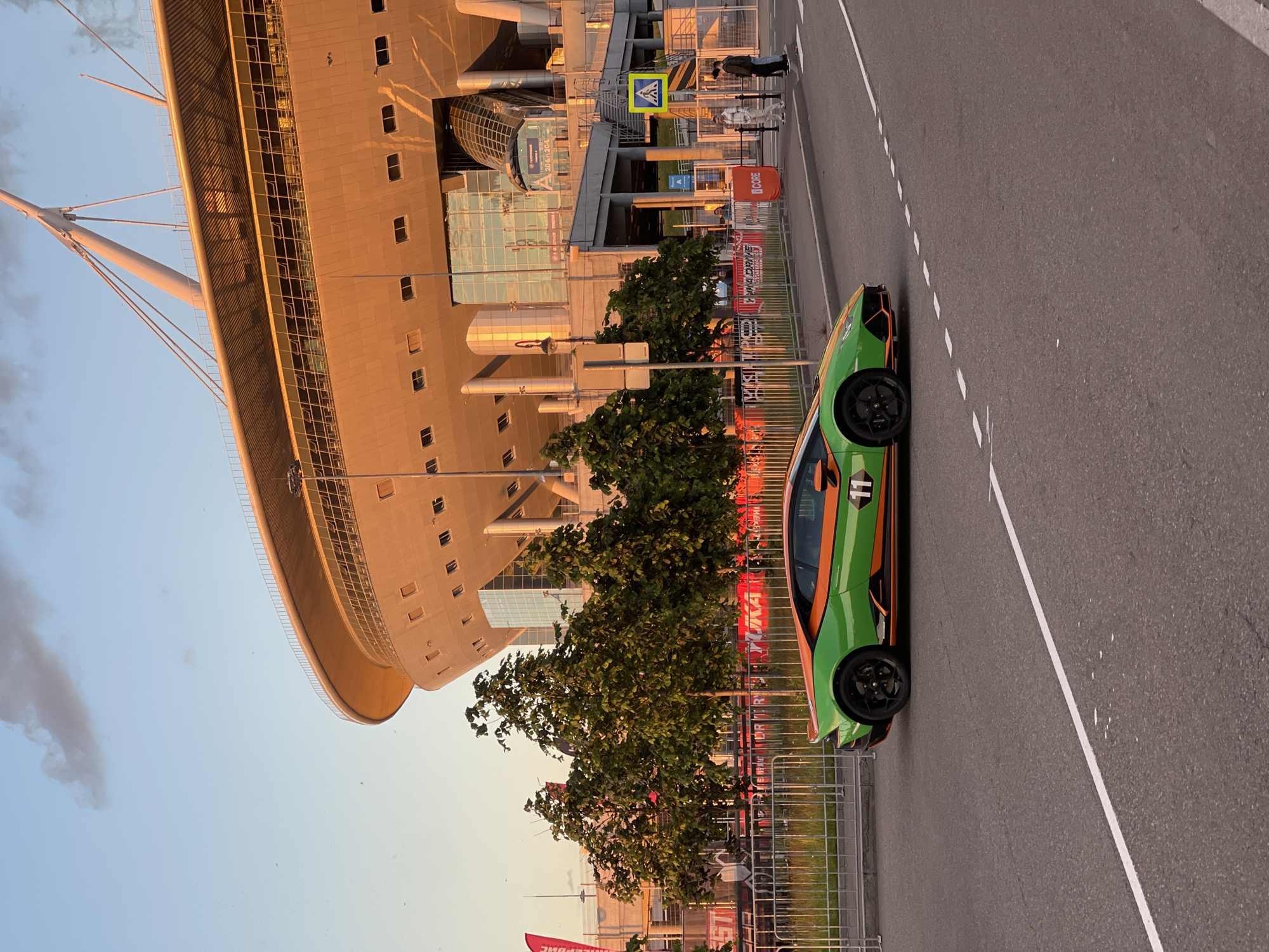 In a city scene at sunset, a vibrant green and orange Lamborghini is parked on an asphalt road. The car, featuring the number "11" on its side, is positioned in the foreground, facing left. Behind the vehicle, a modern stadium with a distinctive curved roof dominates the background. The stadium is illuminated by the warm, golden light of the setting sun, casting long shadows. Several trees with lush green foliage are situated between the road and the stadium, and a metal fence runs along the edge of the property. A few people are visible near the stadium entrance, and a pedestrian crossing sign is posted on the right side of the image. The sky is a clear blue with a few wispy clouds.