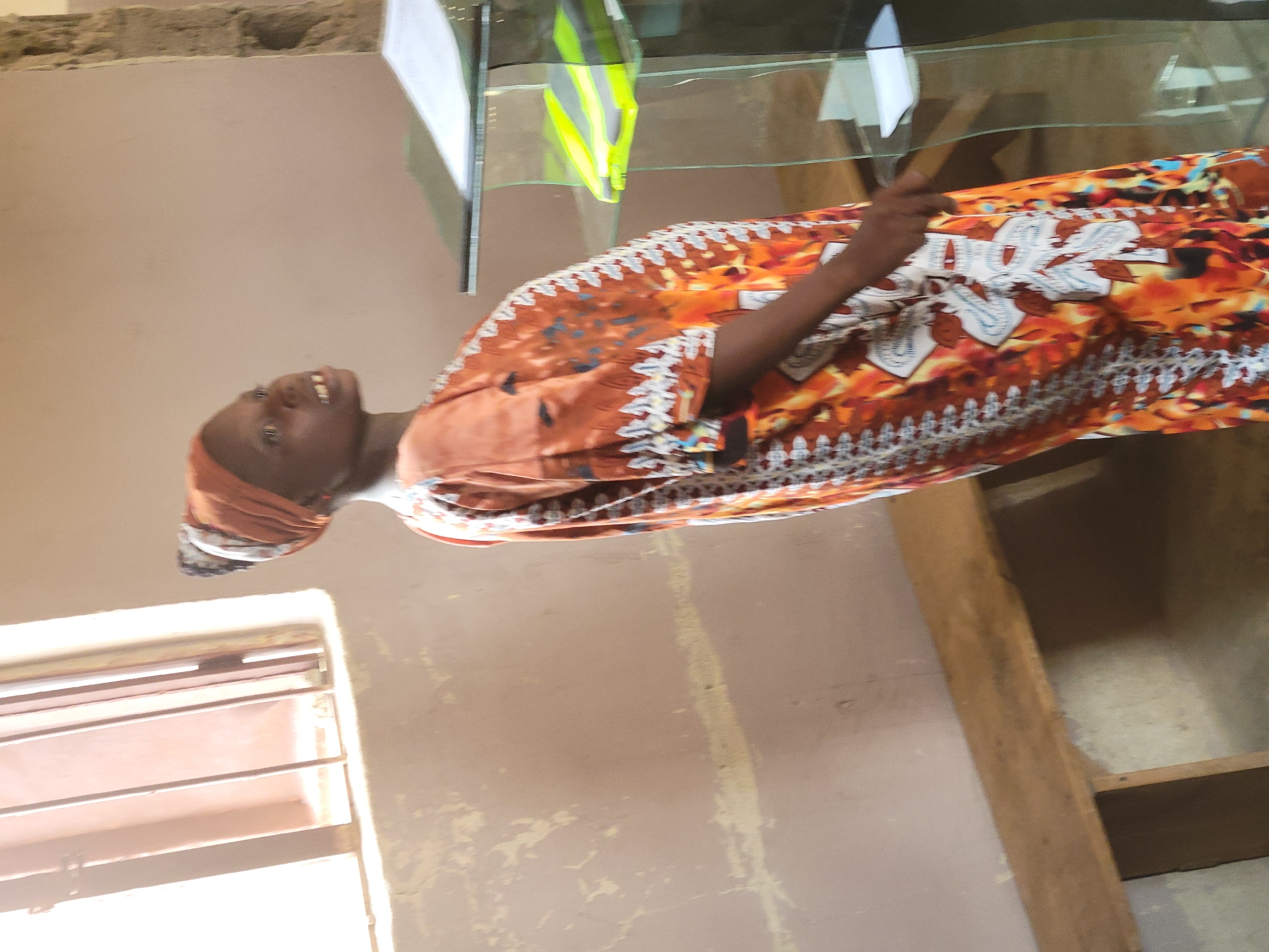 A woman with dark skin and an orange headscarf is standing inside a building in Madalla, Nigeria. She is wearing an orange and white patterned long-sleeved garment. Behind her is a glass display case with various items inside, including a yellow and green object. A window with horizontal bars is visible to the left of the woman. The walls of the building are painted in a neutral color.