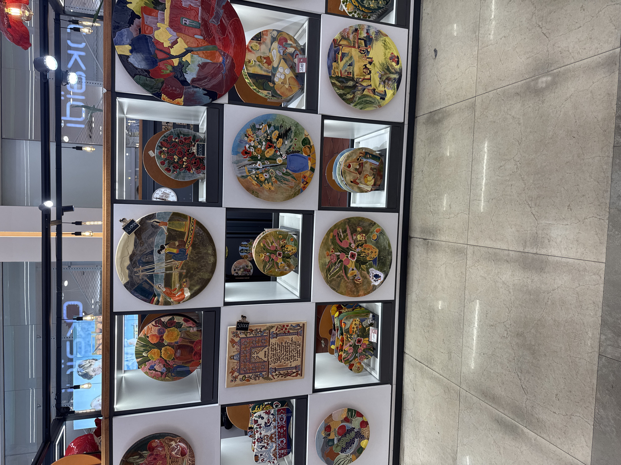 The image shows a retail display of decorative plates and artworks in a store, possibly in Yerevan, Armenia. The display is organized in a grid of illuminated shelving units. The items include various painted plates with floral designs, landscapes, and cultural motifs. Some items have price tags visible. The store appears to be well-lit, with overhead lighting and spotlights illuminating the merchandise. The flooring is tiled with a light-colored marble pattern. The background shows a glimpse of a ceiling with lighting fixtures and signage, suggesting an indoor commercial space.