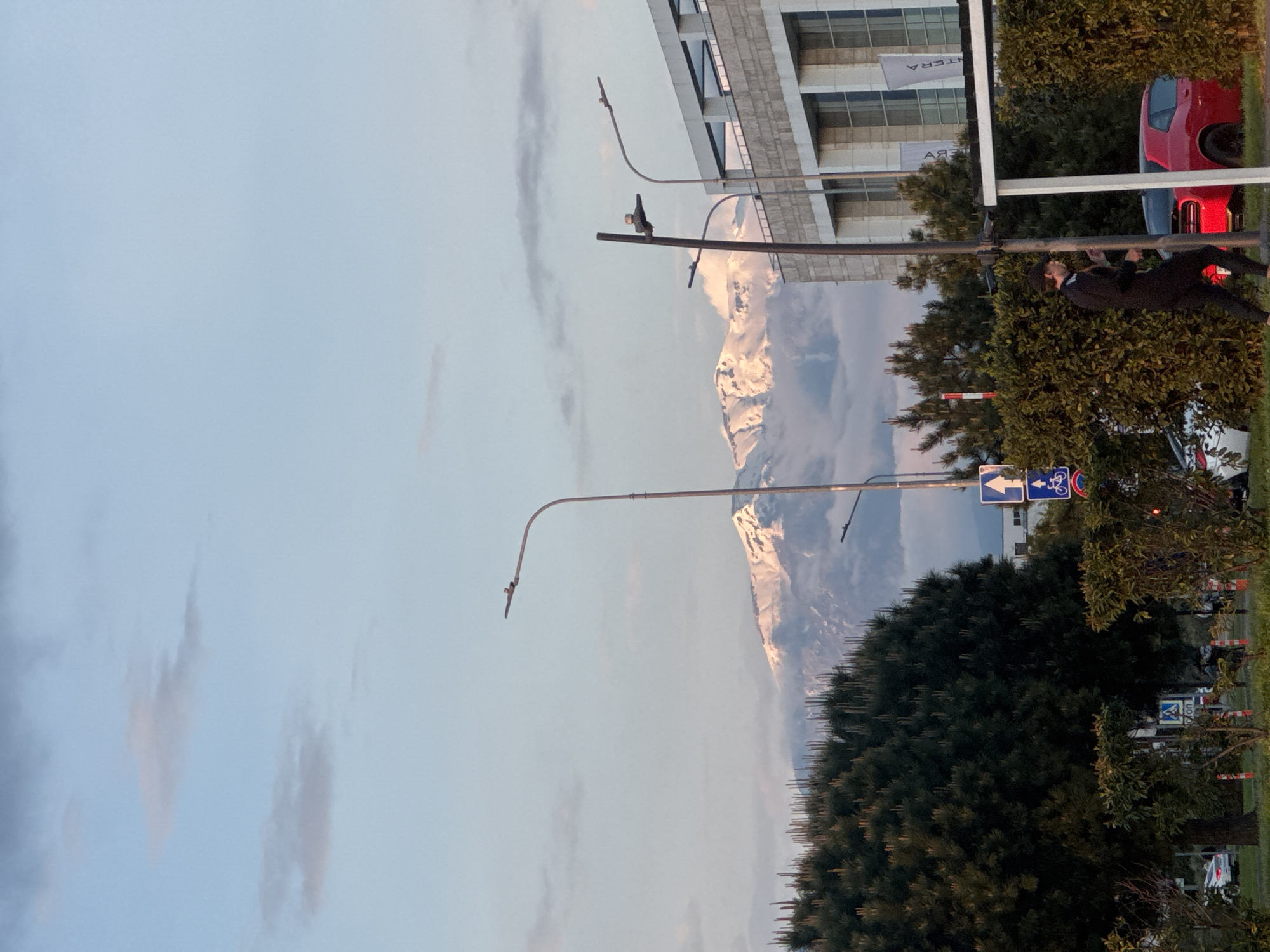 The image shows a street scene in Sirius, Russia, with a view of snow-capped mountains in the background partially obscured by clouds. The foreground features green foliage, including trees and shrubs, and street furniture like lampposts. A road sign indicating direction for pedestrians and cyclists is visible. On the right side, a building with large windows and the word "TERRA" in reflective lettering is present. A red car is parked in front of the building, partially obscured by trees. In the middle ground, a person in dark clothing appears to be hanging from a horizontal structure, possibly engaged in physical activity. A crow is perched on top of the lamppost.