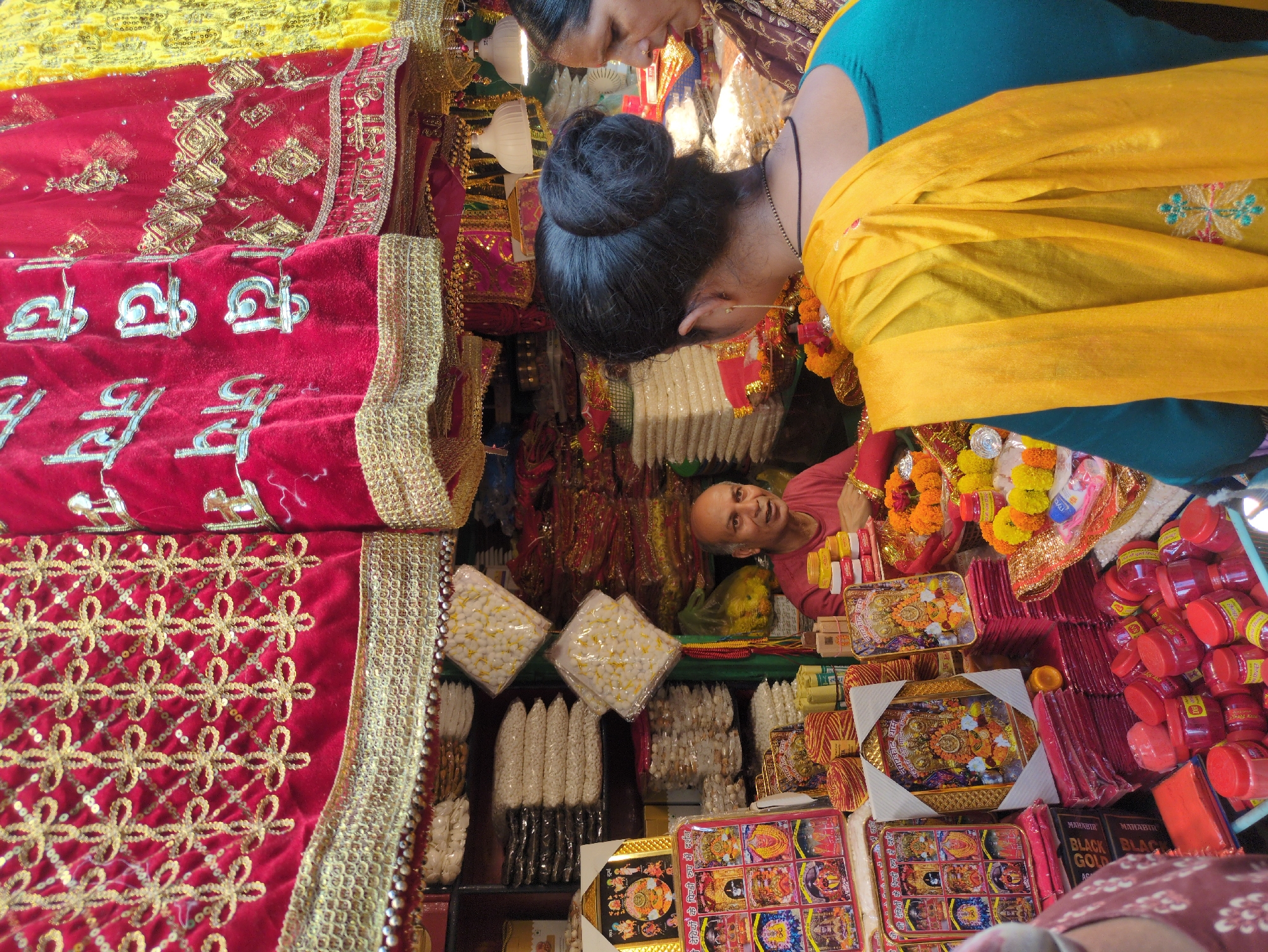 The image depicts a vibrant market stall in Kangra, India, specializing in religious items. In the foreground, a vendor, an older man with a bald head, is seen interacting with customers. He is wearing a reddish-pink t-shirt and appears to be showing them items. Two women are partially visible in the frame, one from behind with her dark hair in a bun, wearing a teal top and a yellow shawl, and another woman to her right, wearing a patterned dark sari.
The stall is laden with various religious paraphernalia. Stacks of red and gold decorative fabrics are displayed prominently, some with intricate patterns and sequins. Piles of packaged white items, possibly religious offerings or sweets, are stacked on shelves behind the vendor. Small framed pictures and decorative items, likely depicting deities, are arranged on display. Red cylindrical containers, possibly containing vermilion powder or other religious substances, are abundant in the lower portion of the image, some in a blue plastic tray. Garlands of marigolds and other flowers are also present, suggesting an active spiritual or festive atmosphere. The overall impression is of a bustling marketplace catering to devotional needs, with bright colors and a sense of commerce intertwined with religious practice.