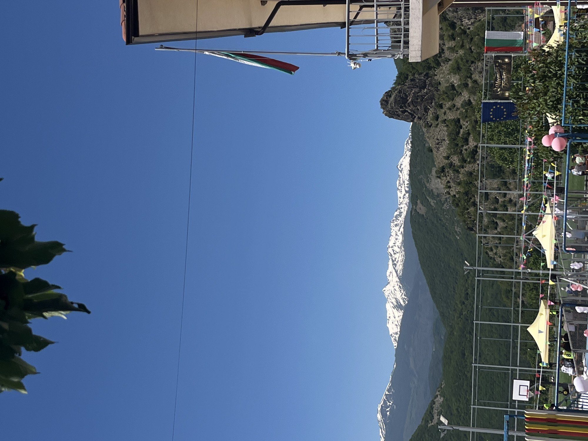 The image depicts a scene in Bukovo, Bulgaria, featuring a building with a Bulgarian flag and European Union flag hanging from it. A mountainous landscape serves as the backdrop, with snow-capped peaks visible. A sports facility, possibly a basketball court, is present with tents and decorative flags. No people are clearly visible.