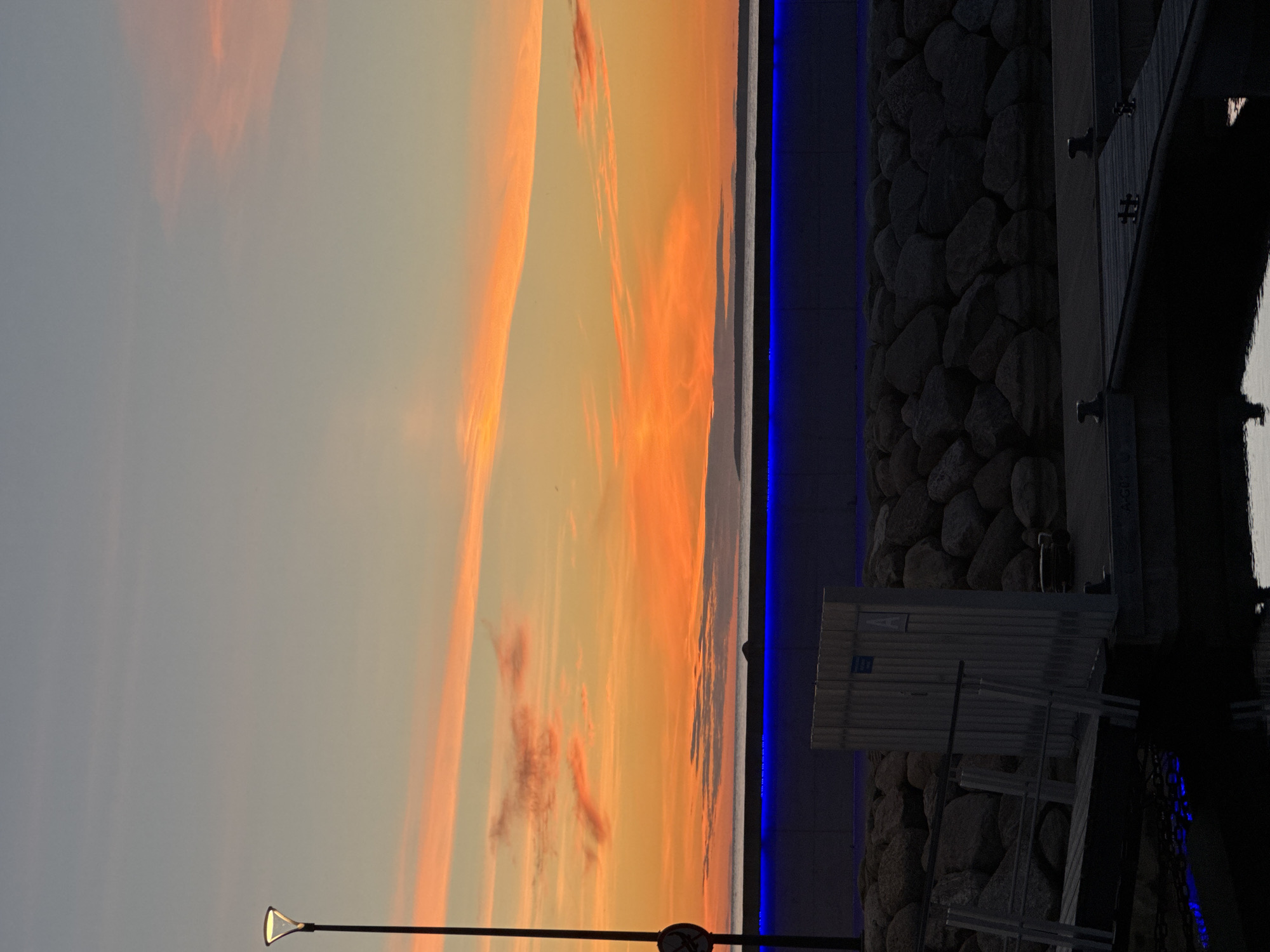 The image depicts a serene sunset scene at a harbor in Tallinn, Estonia. The sky is painted with vibrant hues of orange and pink, with streaks of clouds adding texture to the horizon. A solitary lamppost stands tall on the left side of the frame, its light casting a warm glow. Below the sky, a dark expanse of water meets a rocky shore, which is bordered by a concrete structure illuminated with a strip of blue light. Wooden docks with metal railings jut out into the water, suggesting a functional and well-maintained harbor. The overall atmosphere is tranquil, with the gentle play of light and color creating a picturesque view. The specific location is likely a modern harbor area, possibly designed for recreational boating or as part of a waterfront development in Tallinn.