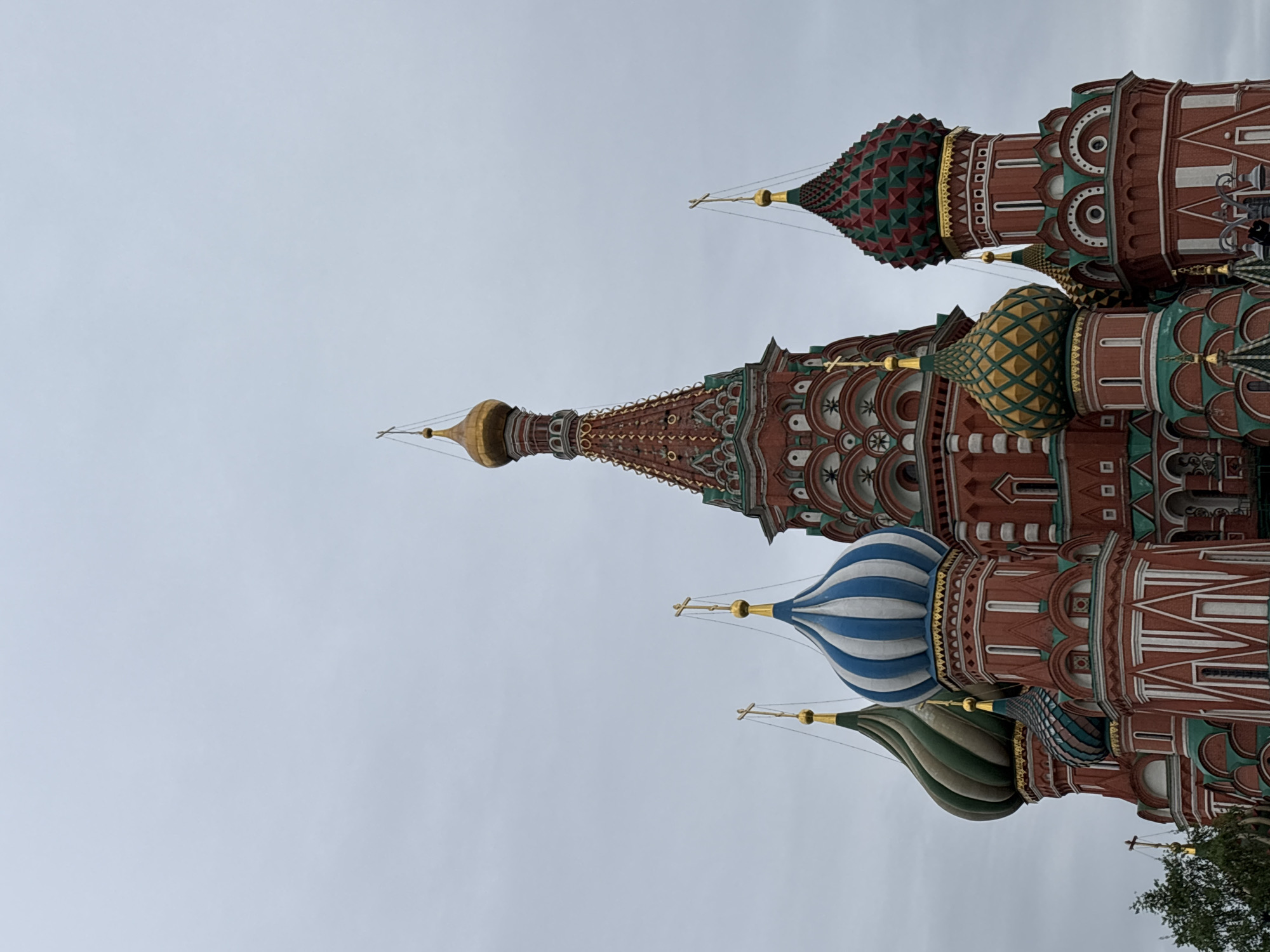 The image shows a partial view of St. Basil's Cathedral in Moscow, Russia, with its distinctive onion domes. The domes are decorated in various colors and patterns, including red, green, gold, blue, and white. The architecture is ornate, featuring intricate brickwork and decorative elements. The sky is overcast.